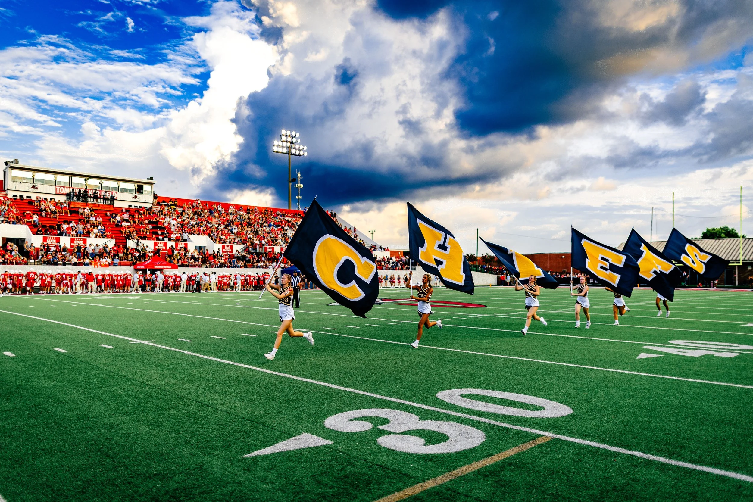 Cheerleaders running across a football field with large flags, crowd in bleachers, cloudy sky overhead.