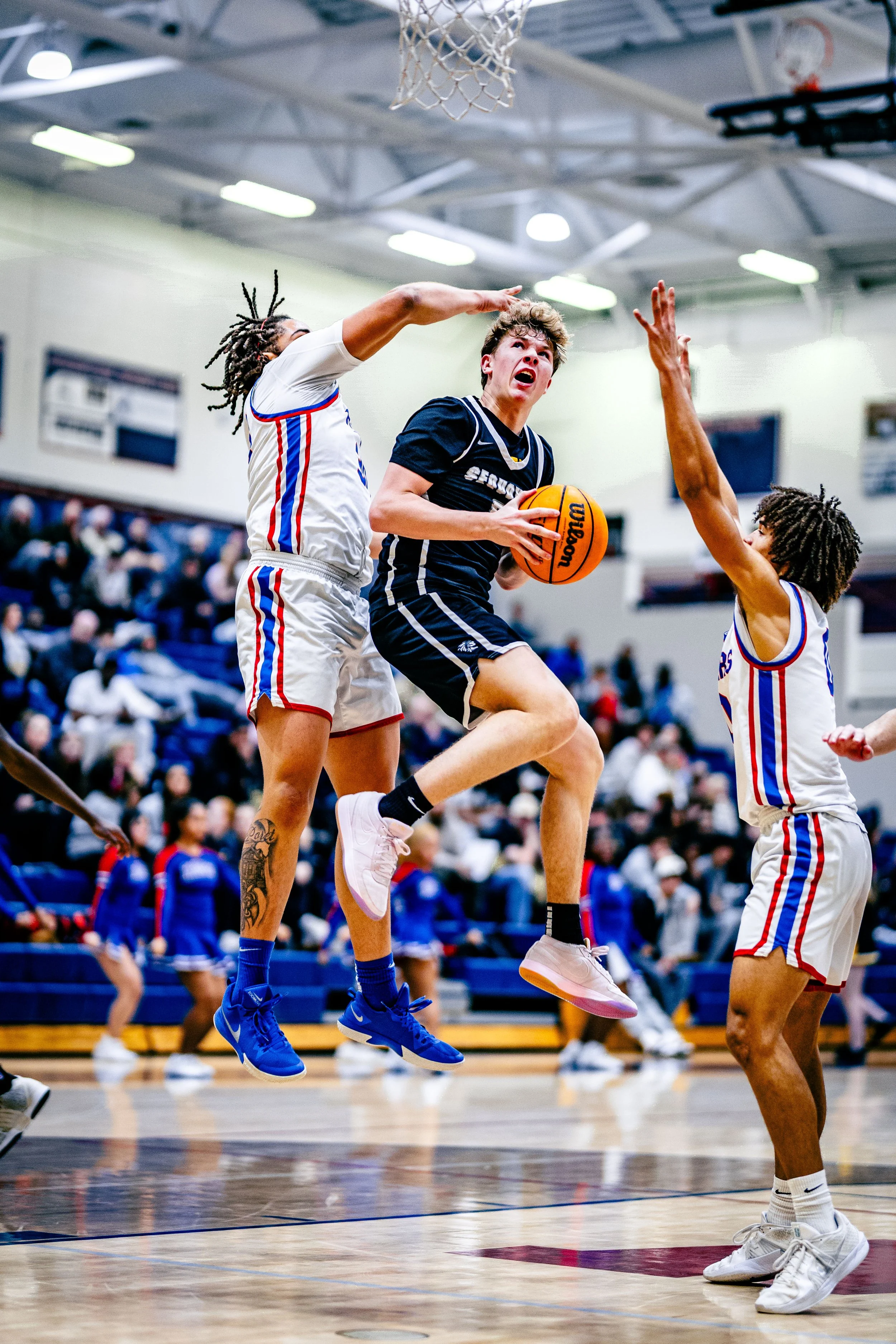 A basketball player in a black jersey jumps with the ball, while two players in white jerseys defend in a gymnasium with spectators.