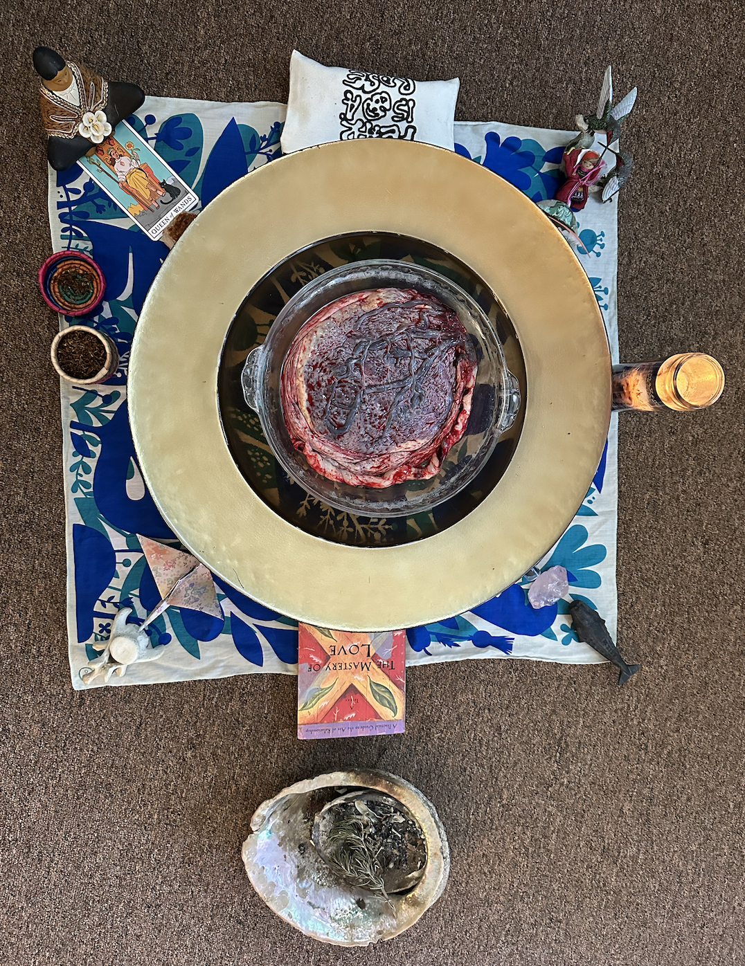Before a placenta encapsulation, the fresh placenta is in the middle of an altar on a circular gold dish. The placenta is surrounded by a candle, The Mastery of Love, an abalone with burning sage, tobacco, cedar, a tarot card, a statue of a mother.