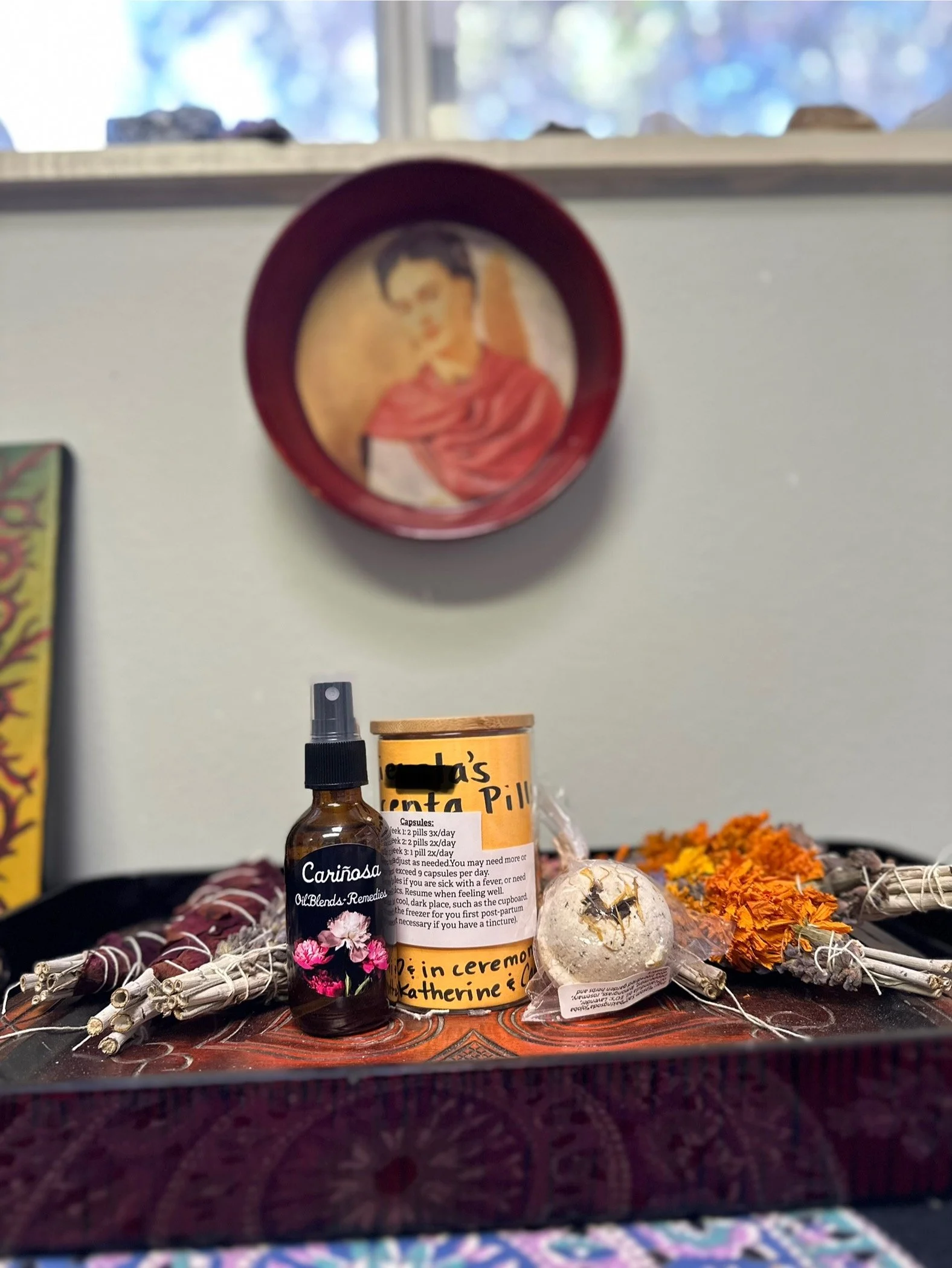 A client’s finished placenta encapsulation in the center with a sage wand and herbal mist to the left and another sage wand and foot scrub to the right. A portrait of Frieda Kahlo is hanging in the background.