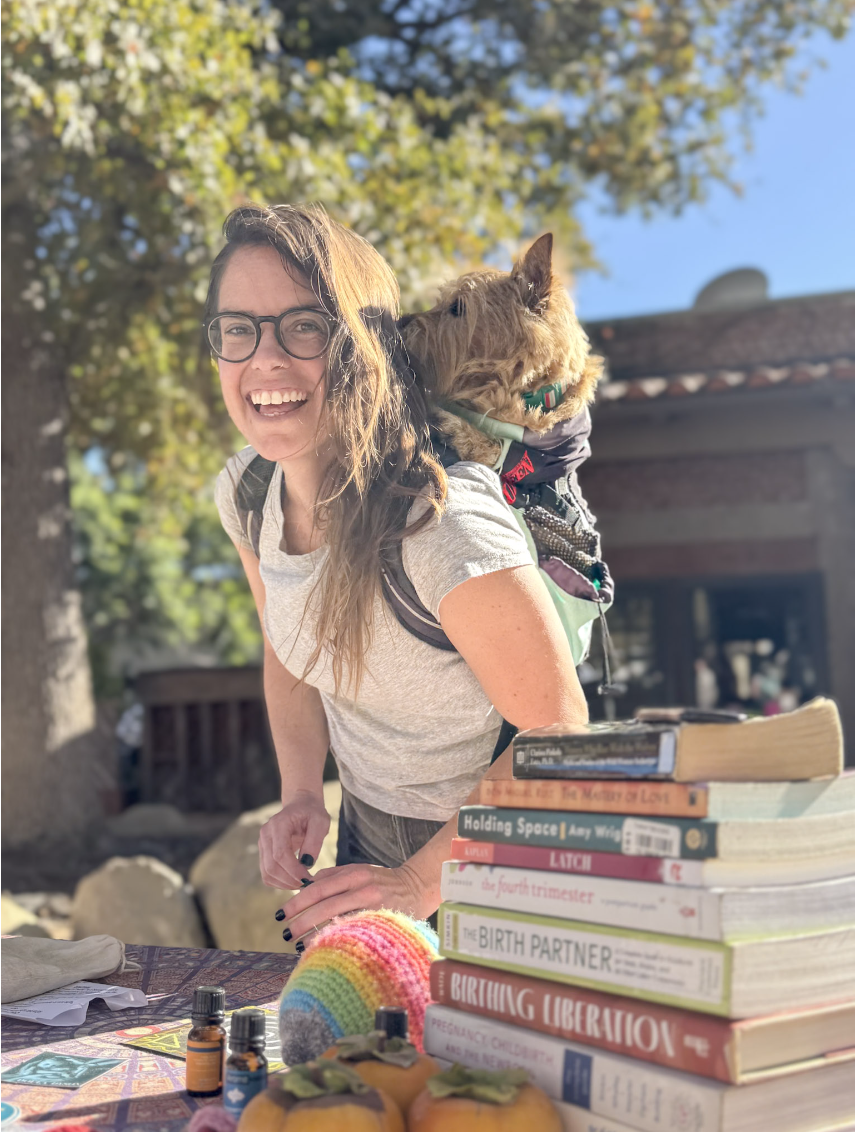 Doula Dyson and her dog Mr. Jones at a Baby and Birth Fair in Ojai California. Doula Dyson with a friendly smile while standing behind a table with birth books and lactation education tools.