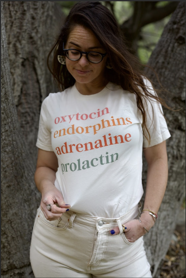 Doula Dyson looking down at a t-shirt she uses as a birth education tool. The t-shirt reads oxytocin, endorphins, adrenaline, and prolactin. She is standing in front of a live oak tree.