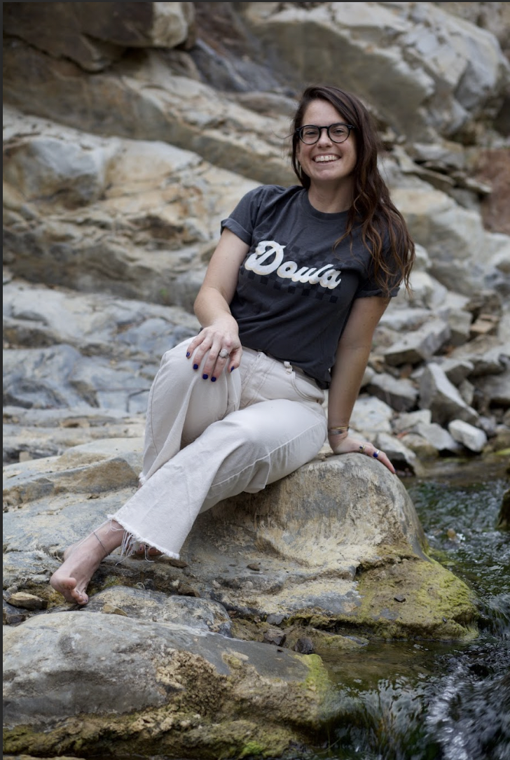 Doula Dyson with friendly smile and a black shirt with doula written in white script casually and confidently sitting on a rock next to a flowing stream.