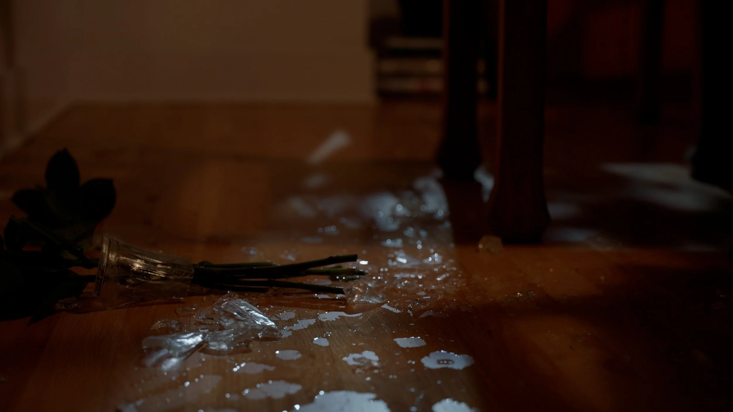 Broken glass vase with spilled water and flower stems on wooden floor.
