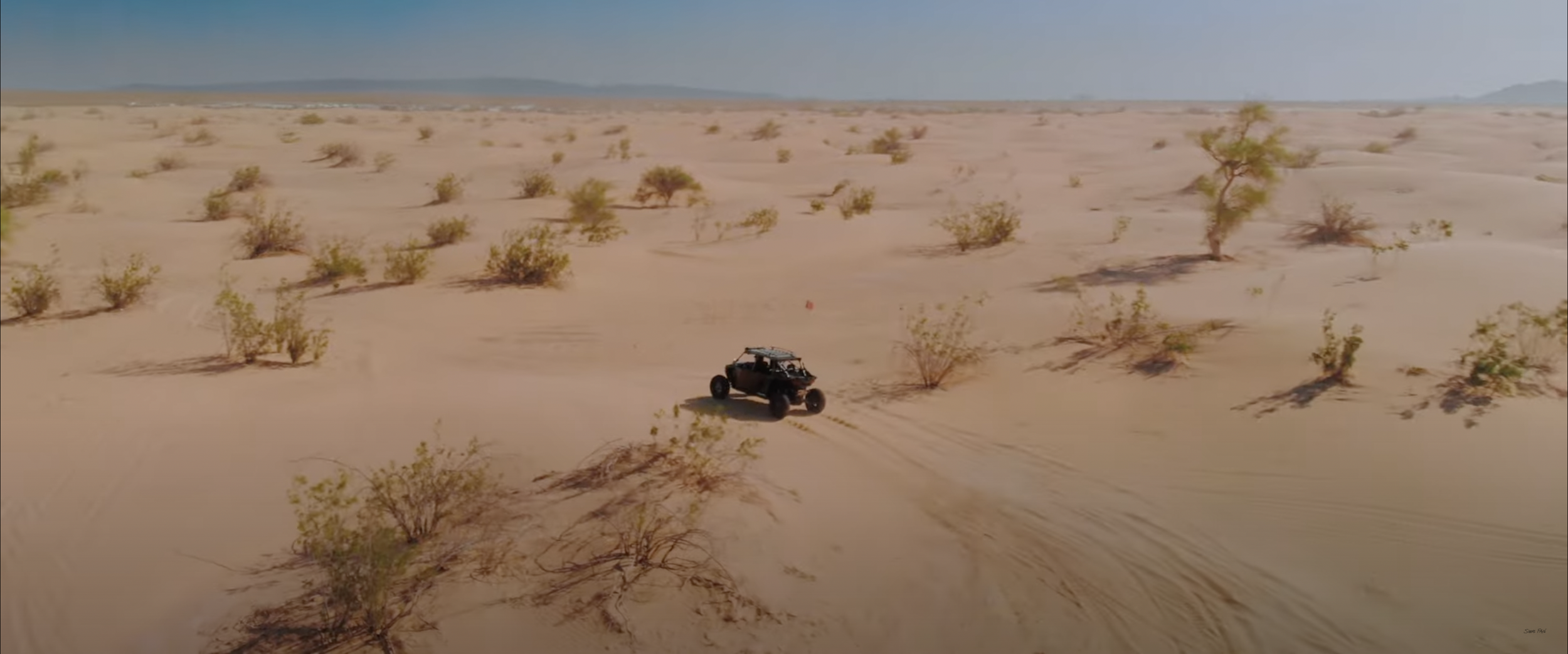 Off-road vehicle driving through desert landscape with sparse vegetation and sand dunes.