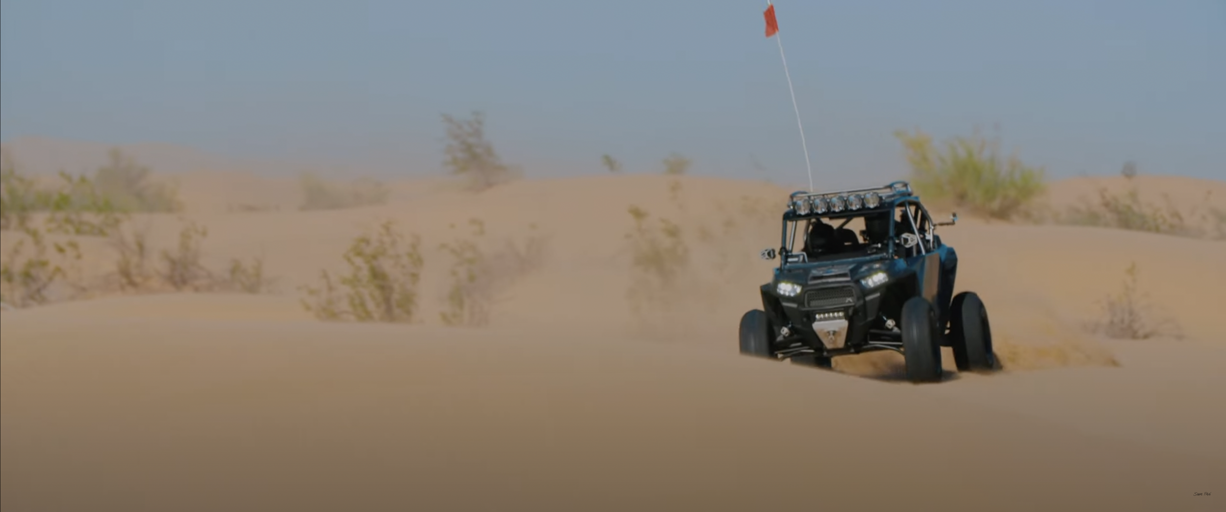 Off-road vehicle driving through sand dunes with a mounted flag.