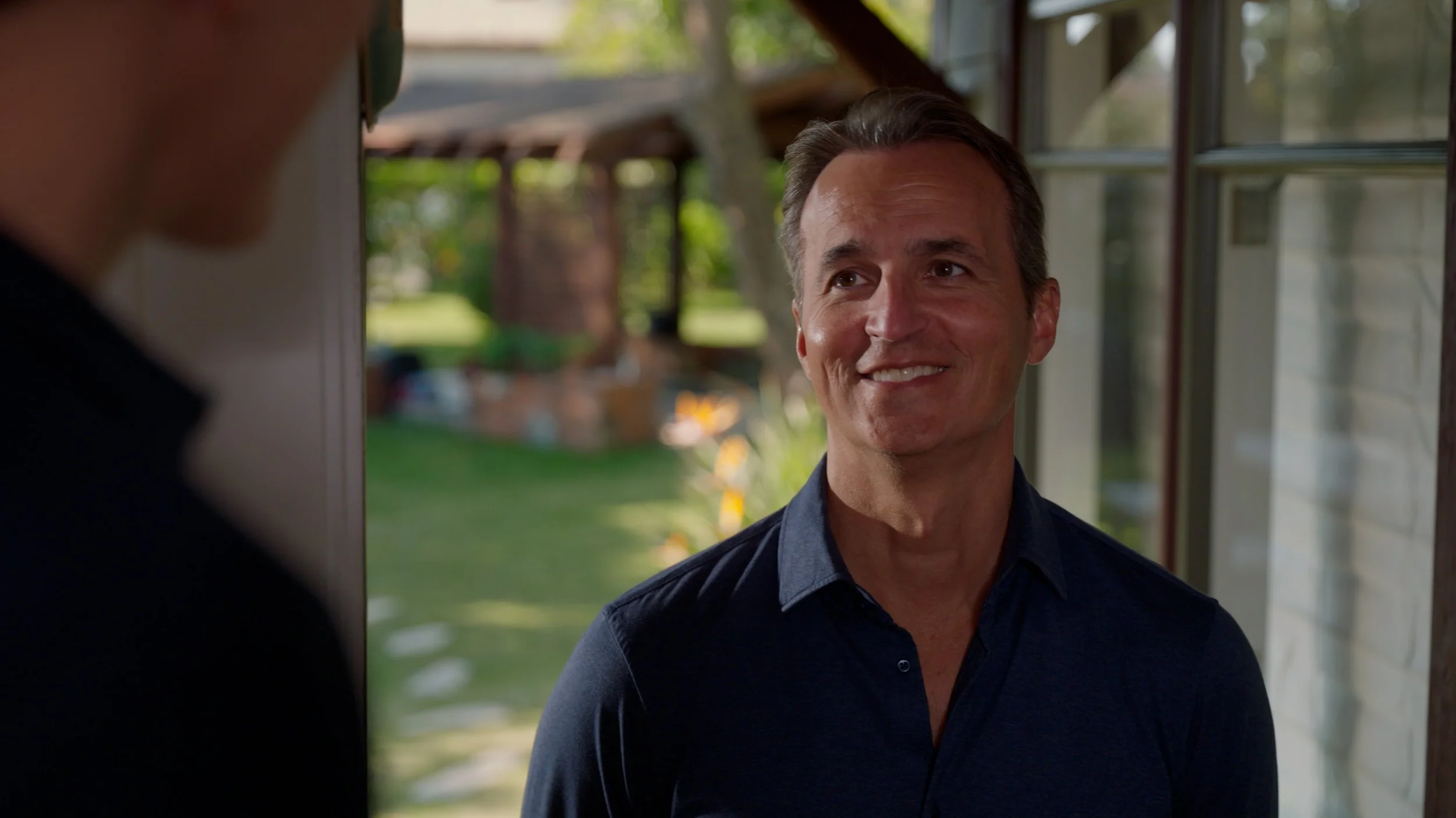 Smiling man in a blue shirt standing near a window with a blurred outdoor background.