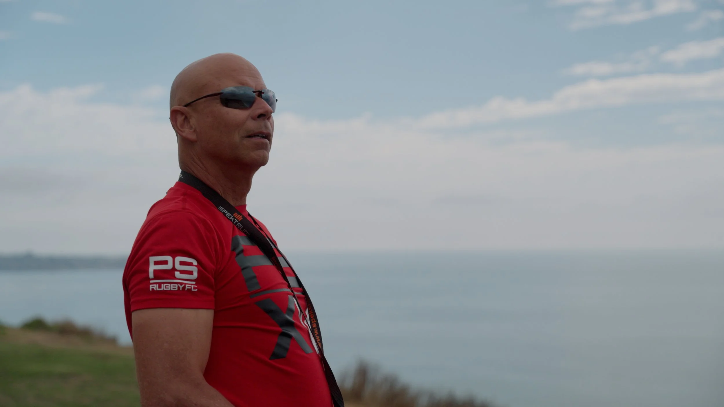 Man in sunglasses wearing a red shirt overlooking the ocean with cloudy sky backdrop.