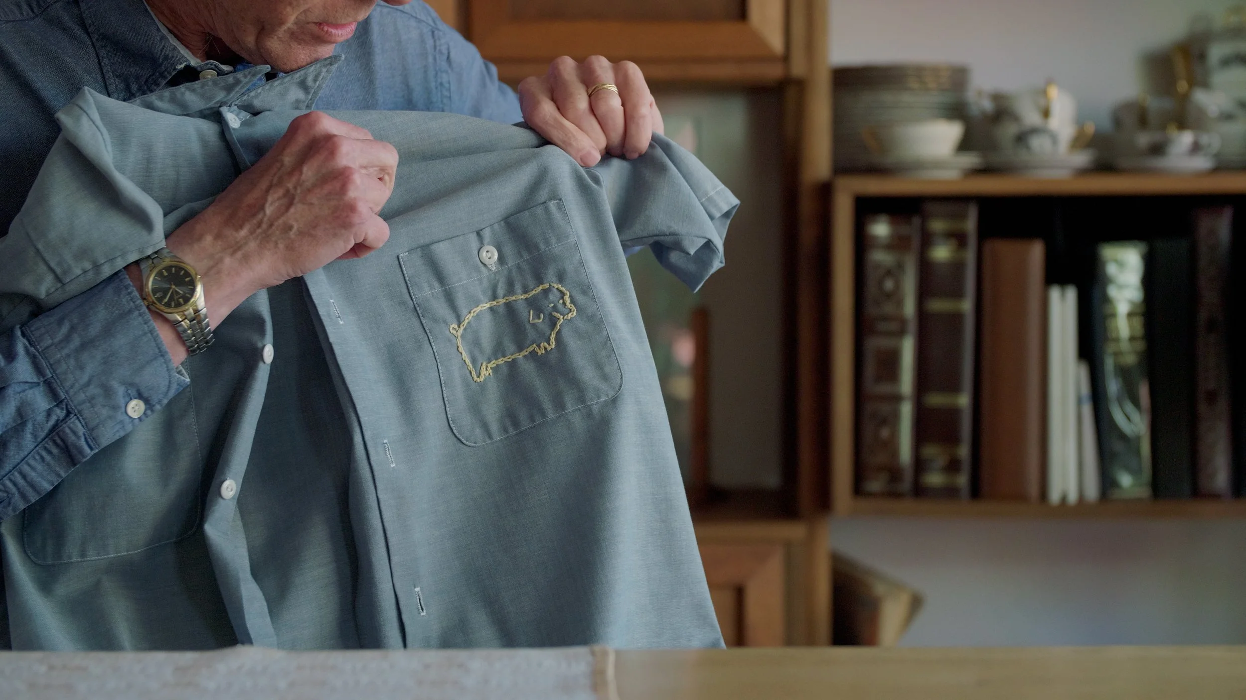 Person holding a grey shirt with embroidered pig design on the pocket, wearing a watch and gold ring, with books and crockery in the background.