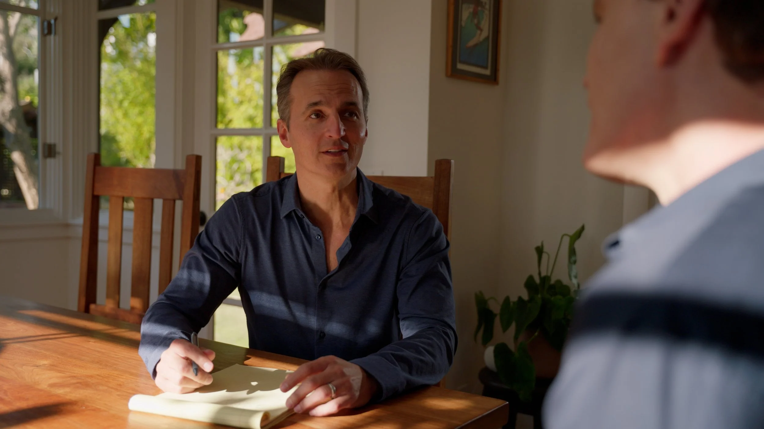 Two people sitting at a wooden table in a sunlit room with a houseplant and a framed picture in the background.