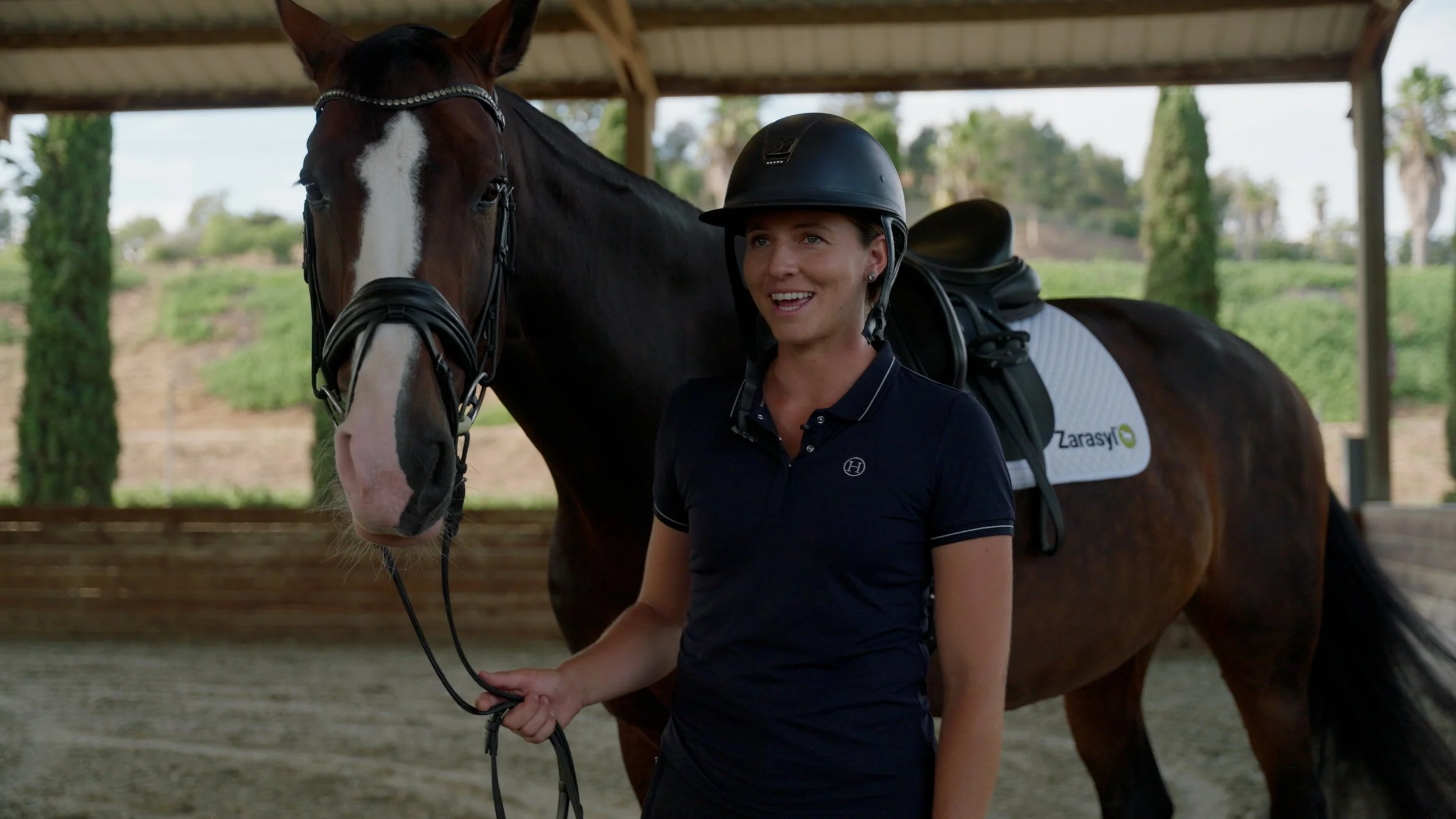 Person in riding gear standing next to a brown horse wearing a bridle and saddle in an outdoor stable area.