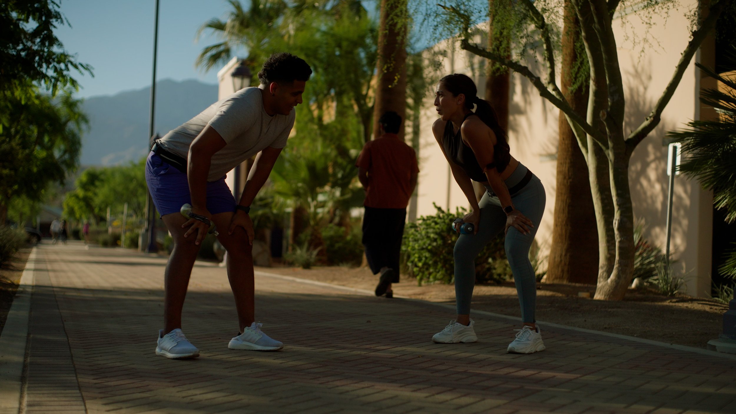 Two people standing outdoors with exercise weights, wearing athletic clothes, under trees.