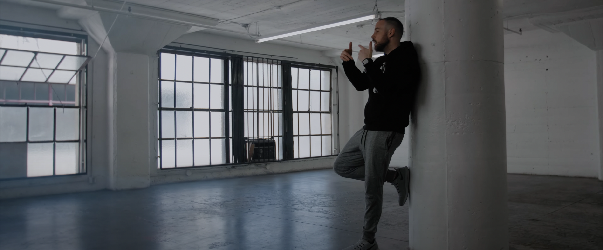 Person in a hoodie leaning against a pillar indoors, gesturing with hands.
