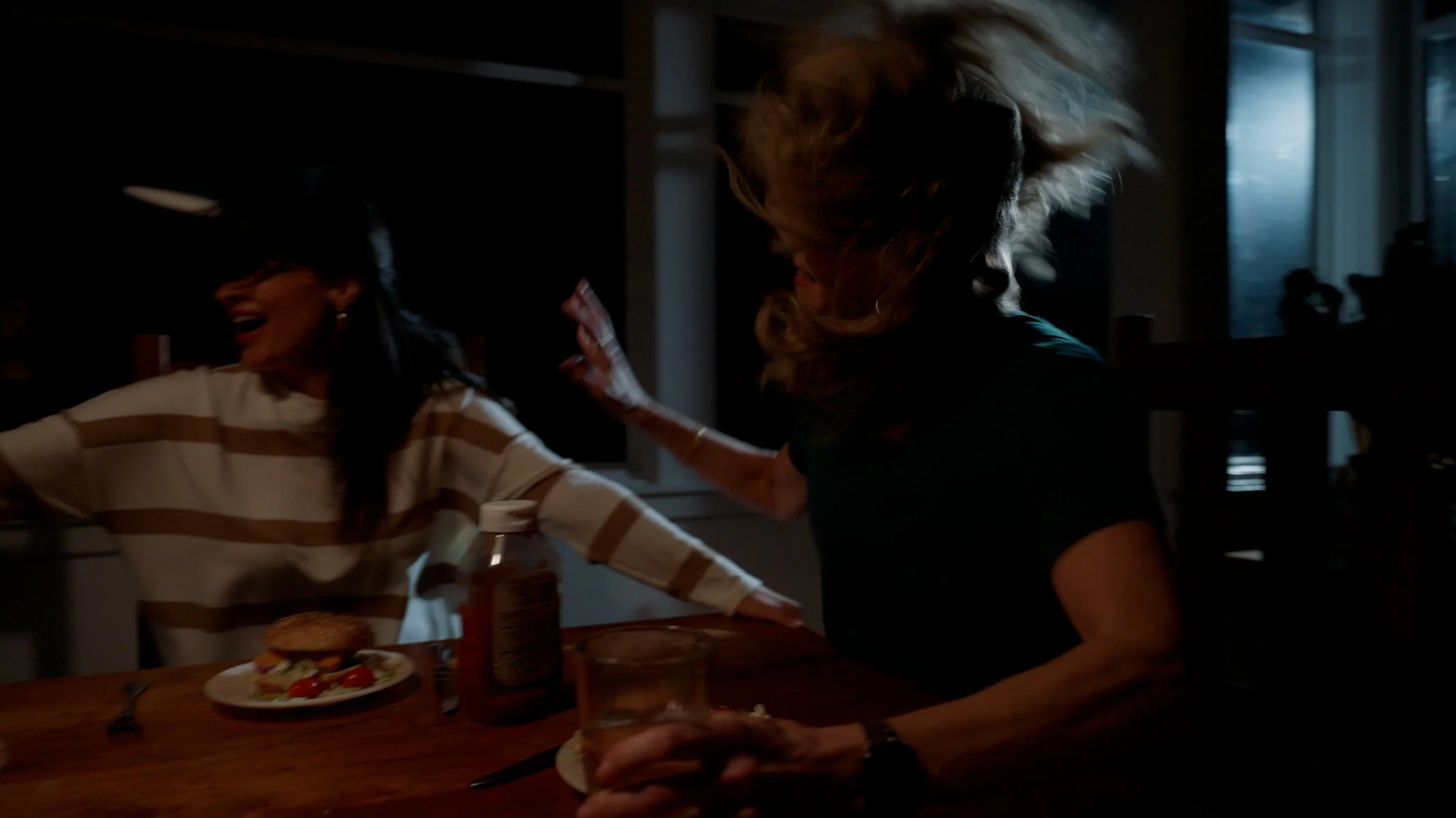 Two women in a dark room with disheveled hair, one laughing, sitting at a wooden table. A burger and tomato salad on a plate, a ketchup bottle, and a glass are visible on the table.