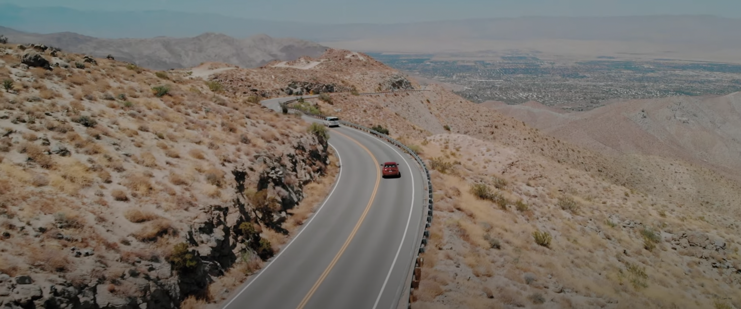A red car driving on a winding desert mountain road with dry brush and distant hills.