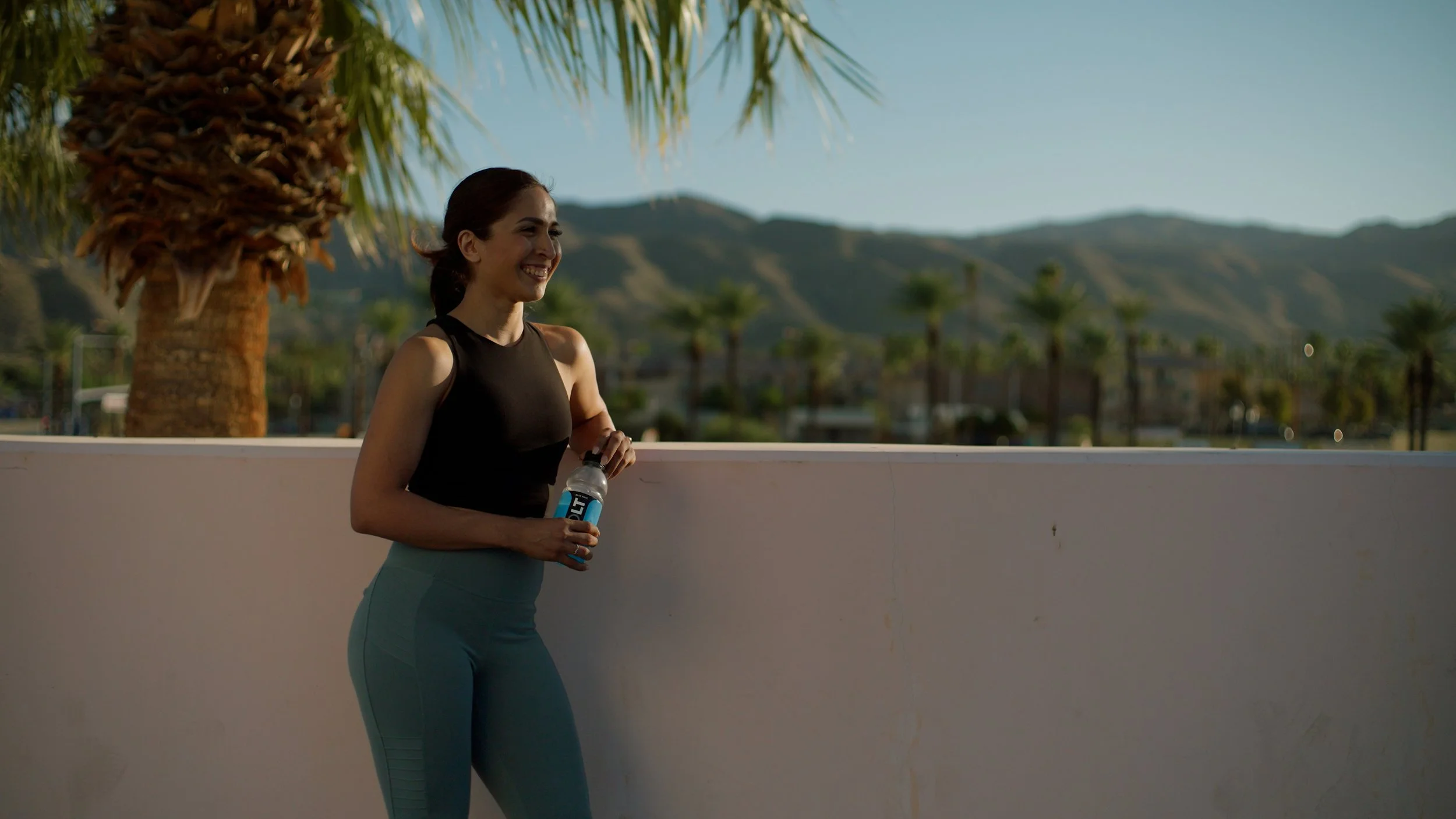 Woman exercising outdoors with water bottle, wearing black top and blue leggings, standing near palm trees and mountains.