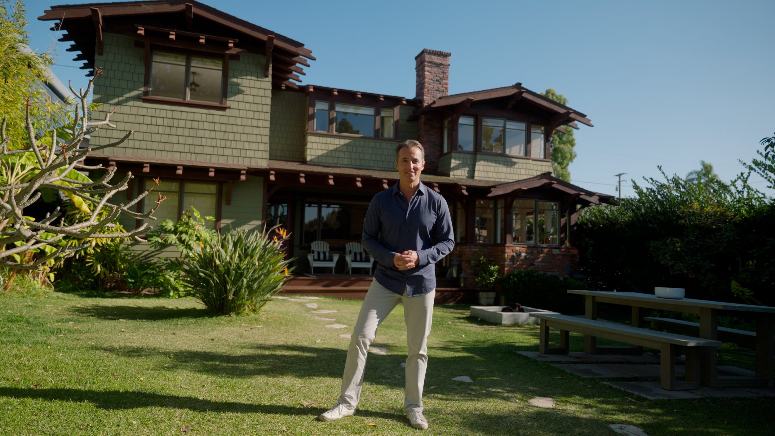 Man standing in front of a two-story green house with wooden accents, surrounded by a garden and lawn, on a sunny day.