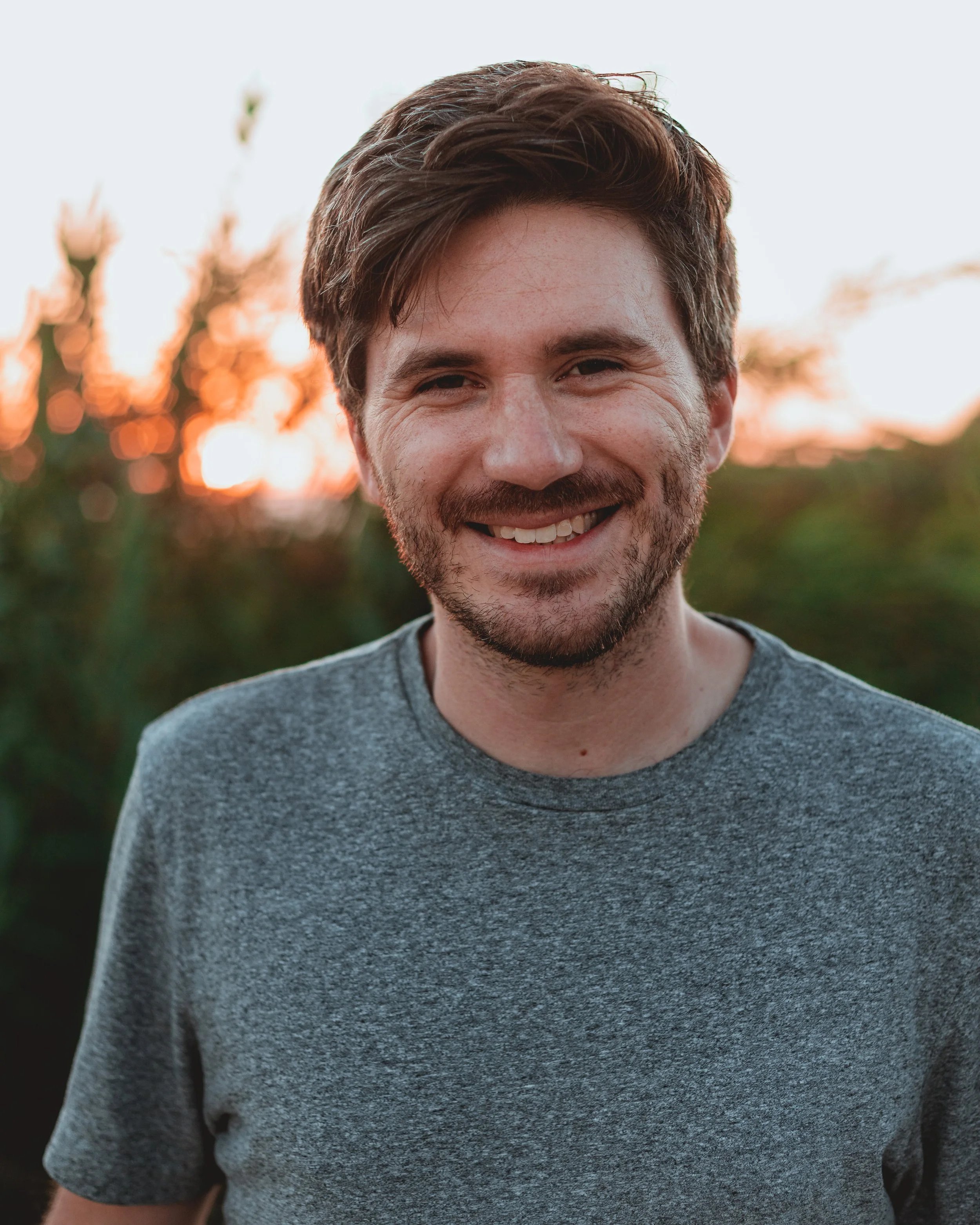 Smiling man in a gray t-shirt with blurred outdoor background at sunset.