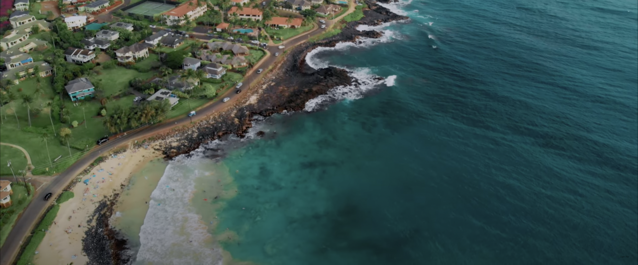 Aerial view of a coastal neighborhood with houses, a road, rocky shoreline, and ocean waves.