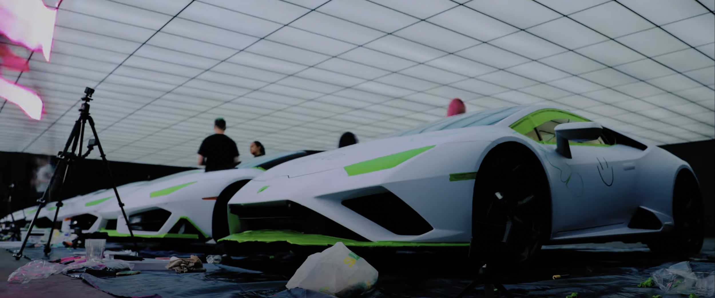 Two white sports cars with green highlights in a brightly lit room with people and equipment around.