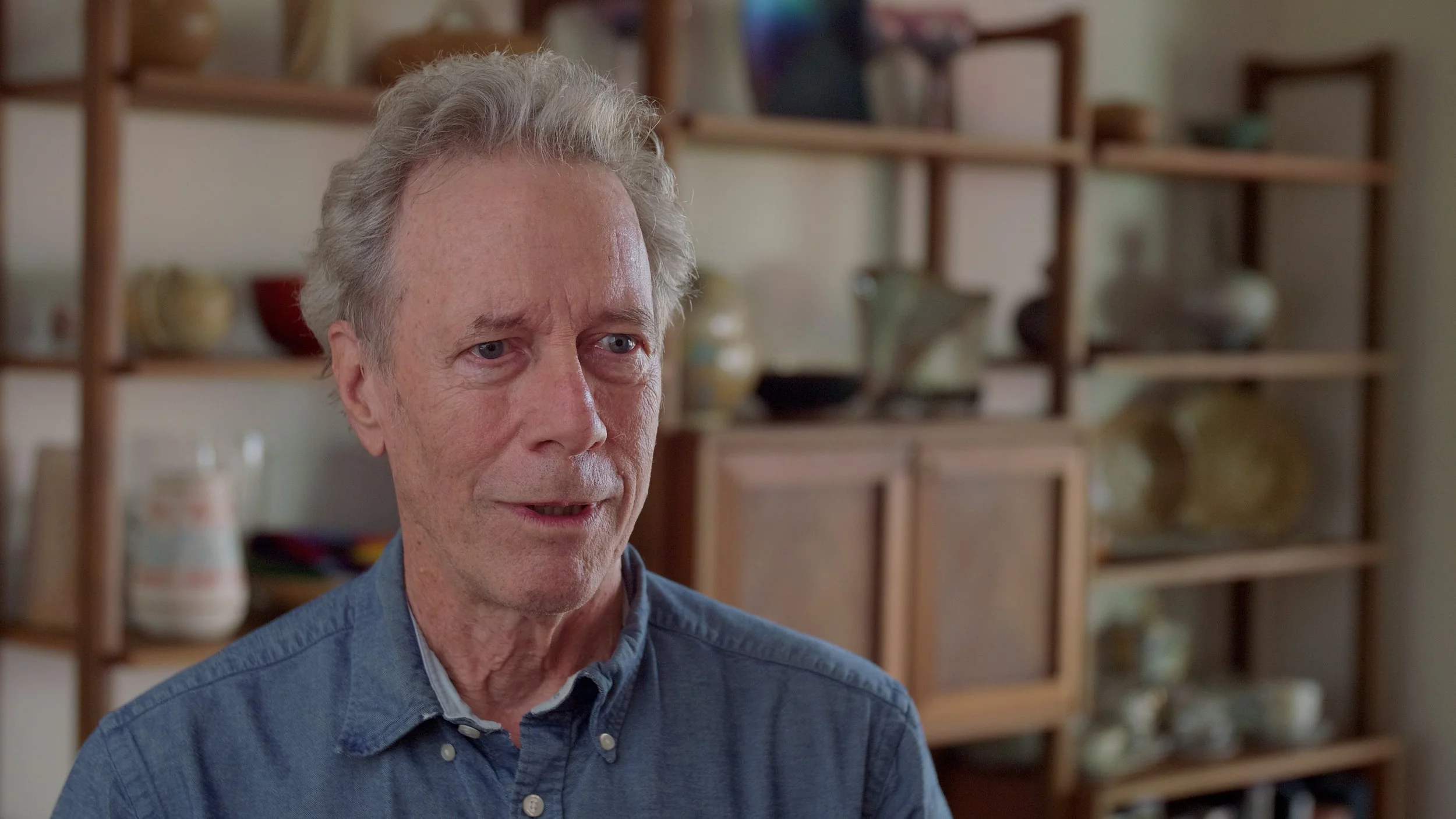 Elderly man with gray hair wearing a blue shirt in front of a wooden shelf.