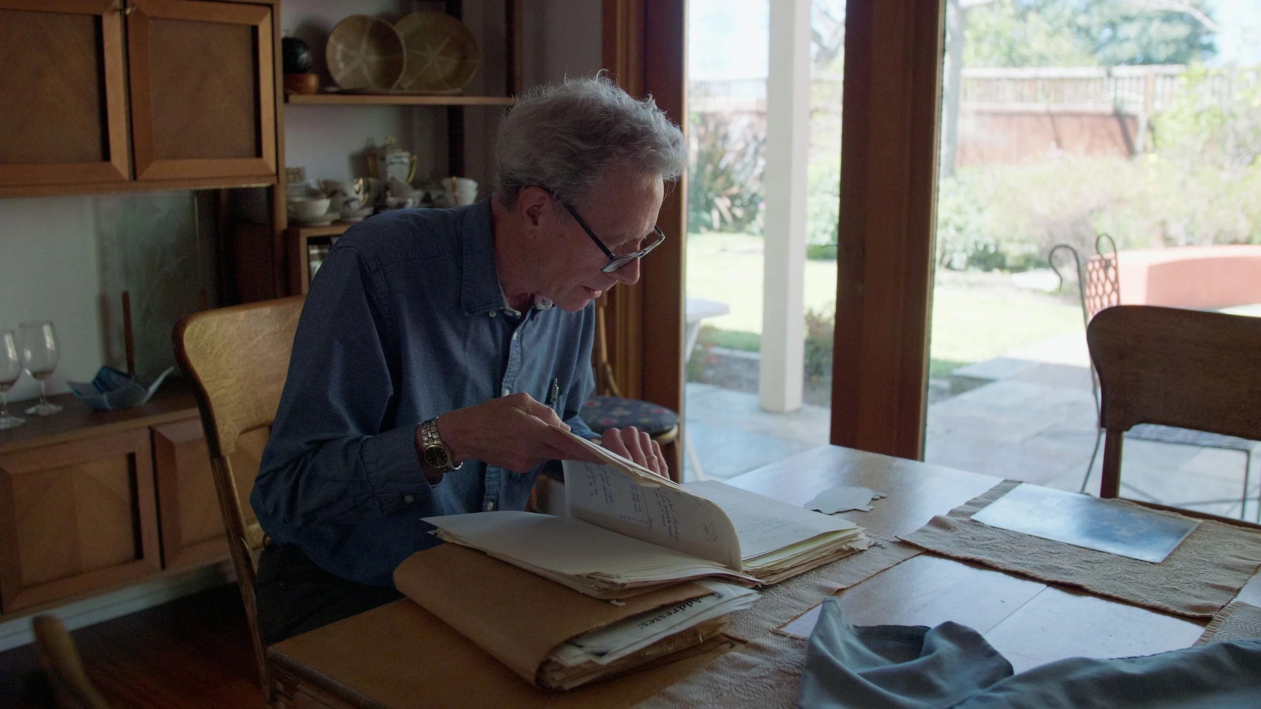 Man reading documents at a wooden table near a large window with garden view.