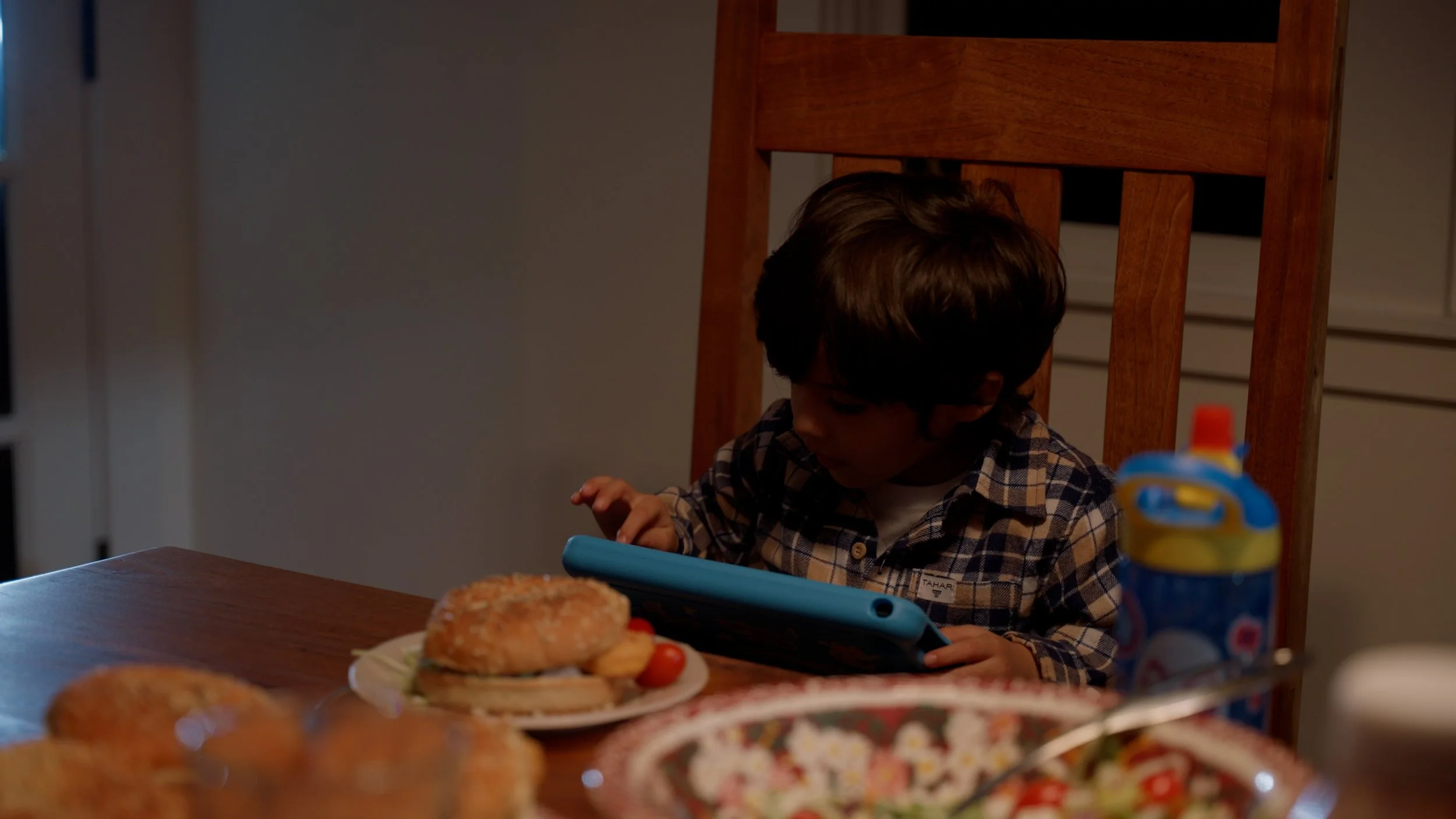 Child using tablet at dining table with food including sandwich and salad.