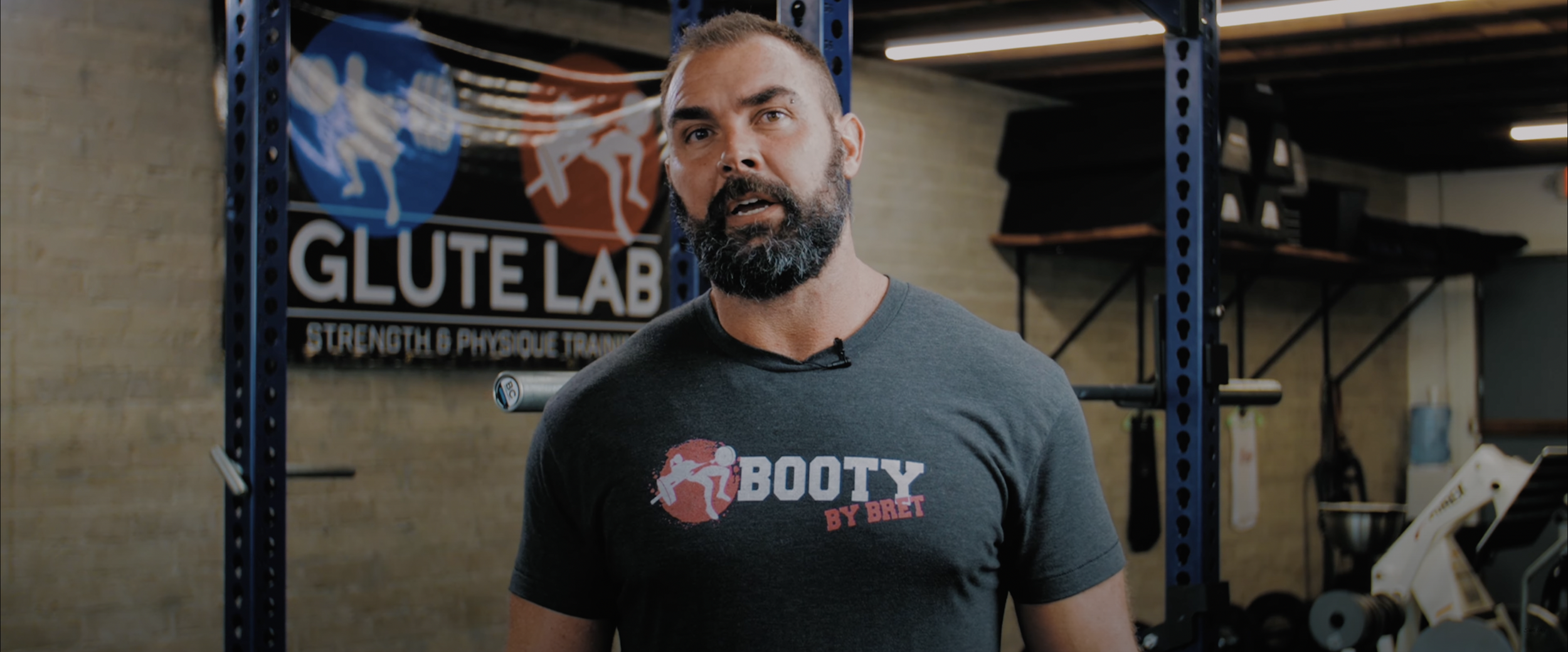 Man in a gym wearing a "Booty by Bret" shirt with "Glute Lab" sign in background.