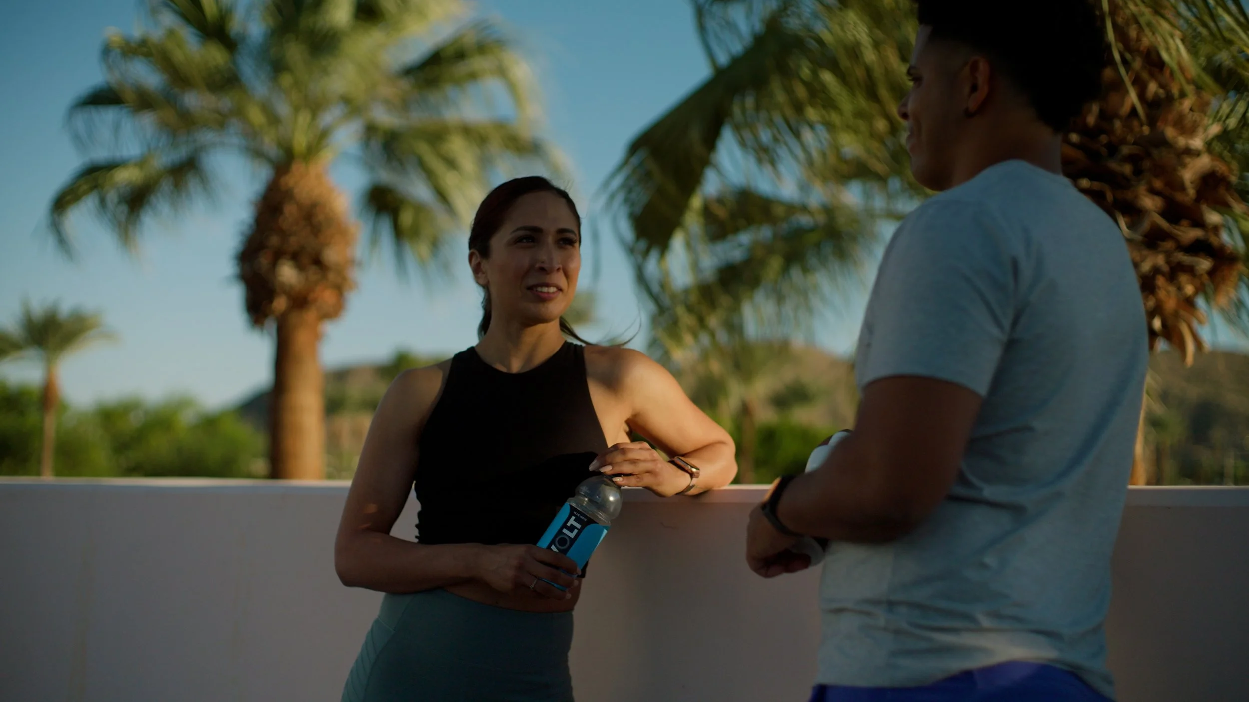 Two people talking outdoors, one holding a water bottle, with palm trees in the background.