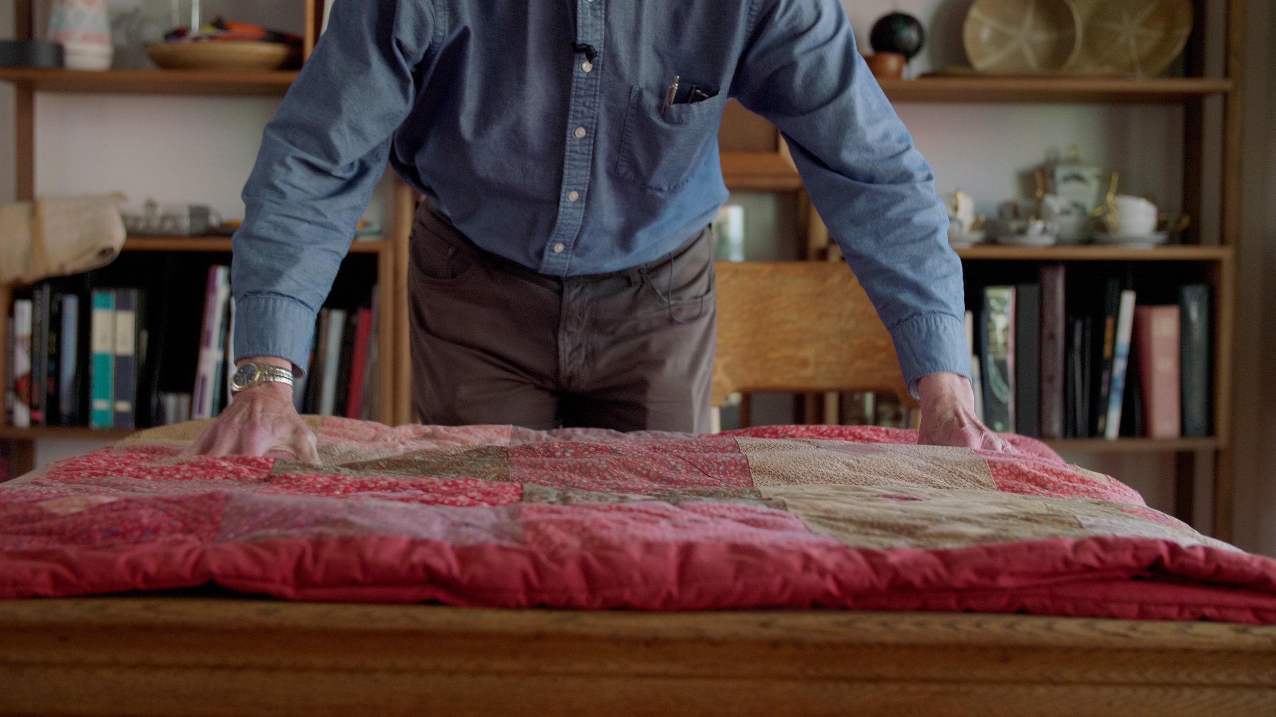 Person arranging quilt on wooden table, bookshelf in background.