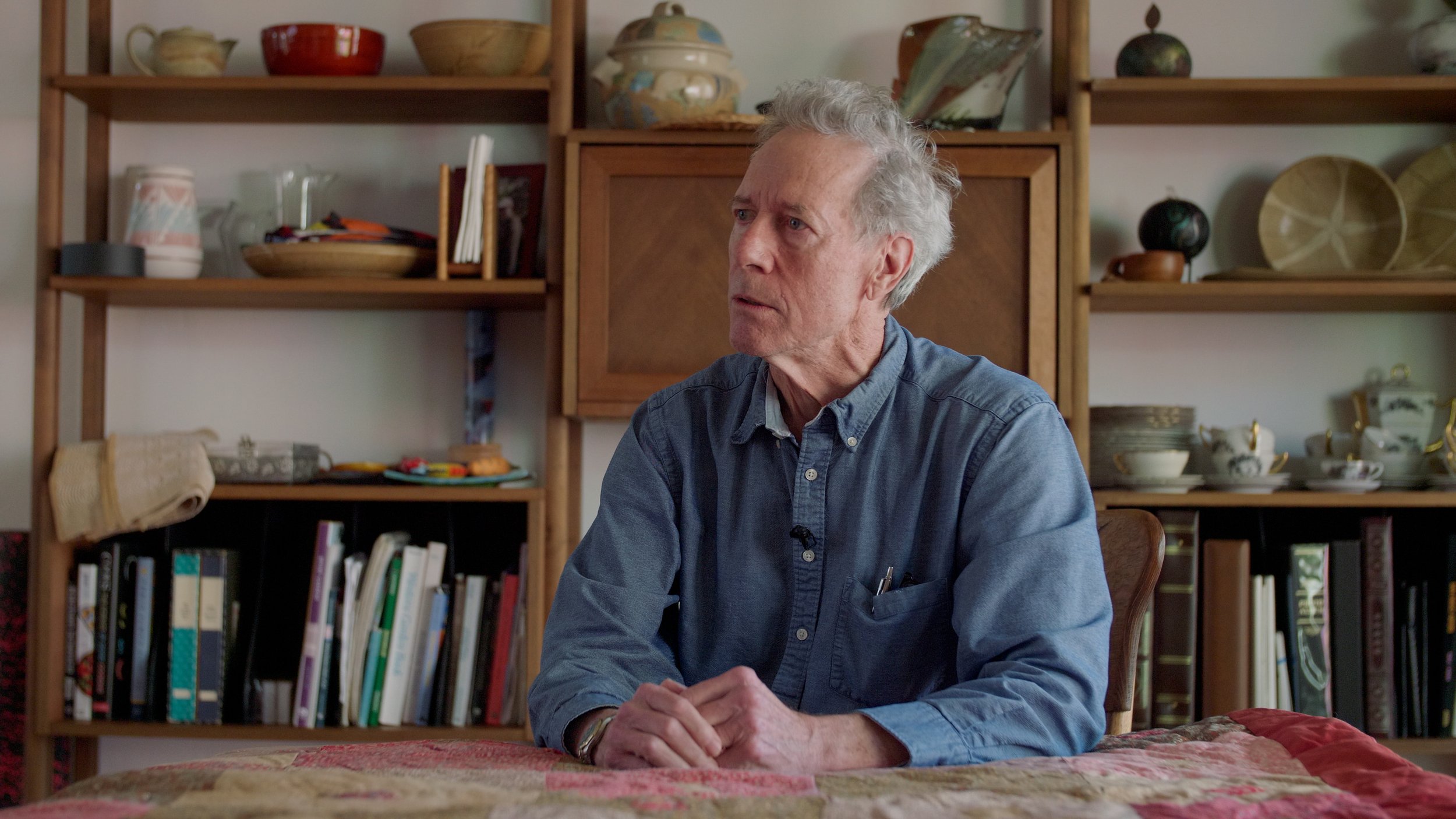 Older man sitting at a table in a room with wooden shelves filled with books and pottery.