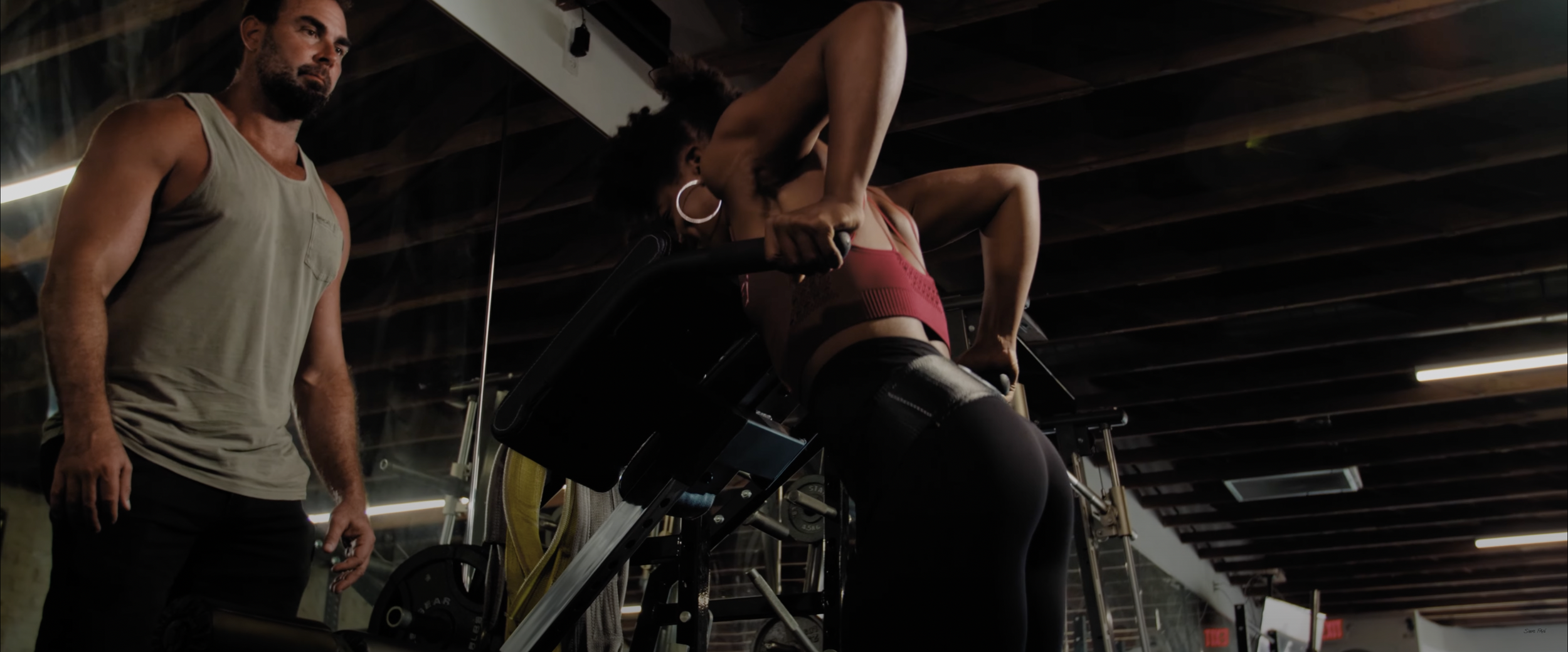 Person lifting weights on a Smith machine in a gym with a trainer observing.