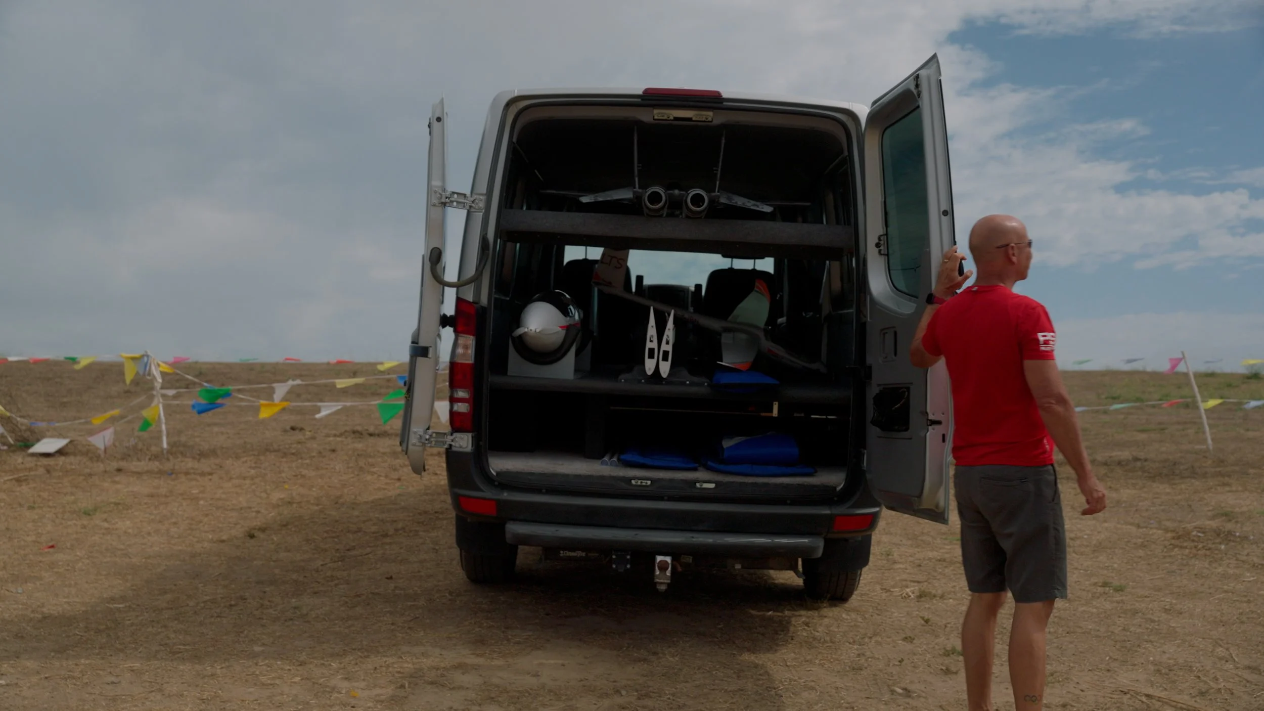 Rear view of an open van with a model aircraft inside, parked on a dirt field with colorful flags. A person in a red shirt stands by the van.