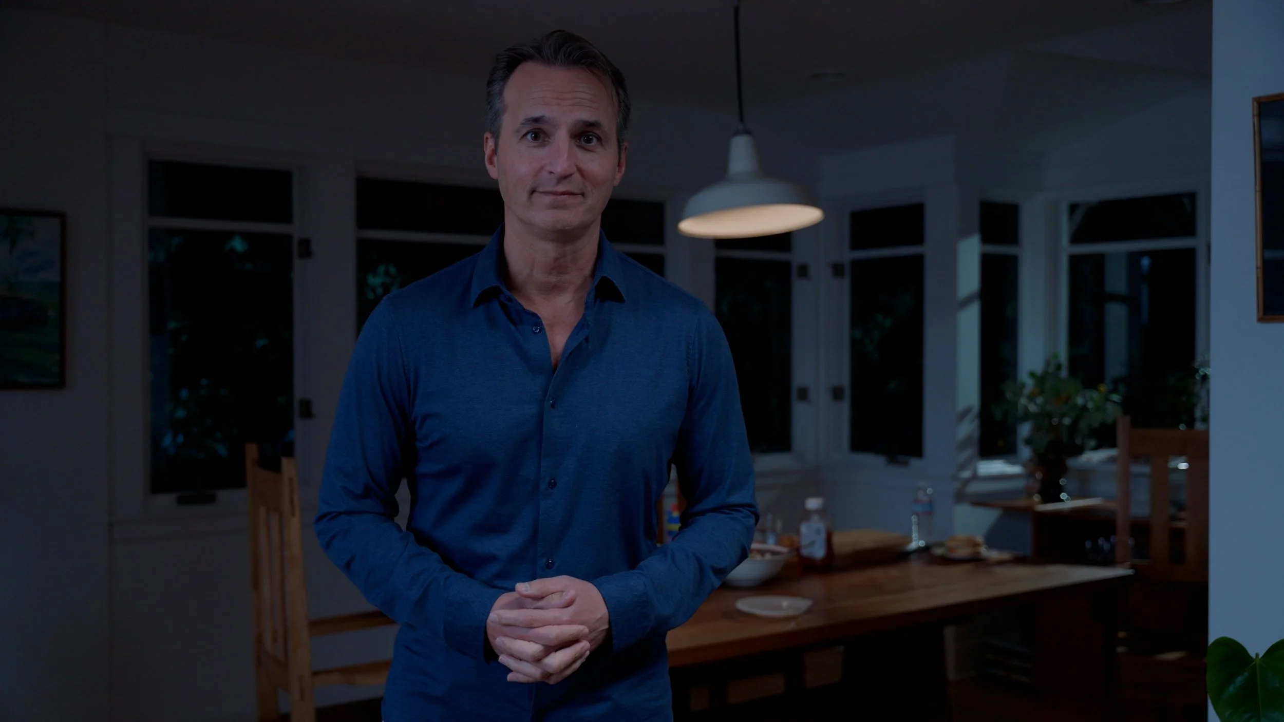 Man in blue shirt standing in dimly lit kitchen with wooden table and chairs, window in background.