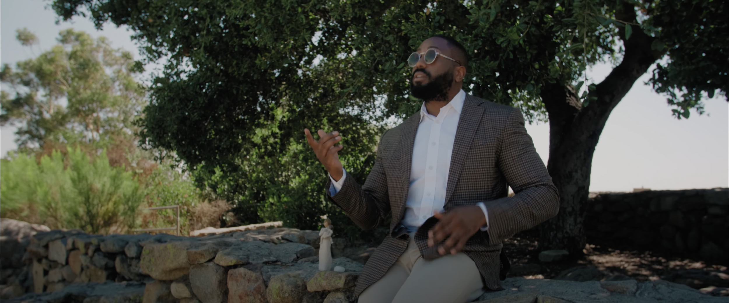 Man in sunglasses and a suit sitting on a stone wall outdoors, near a tree and greenery.