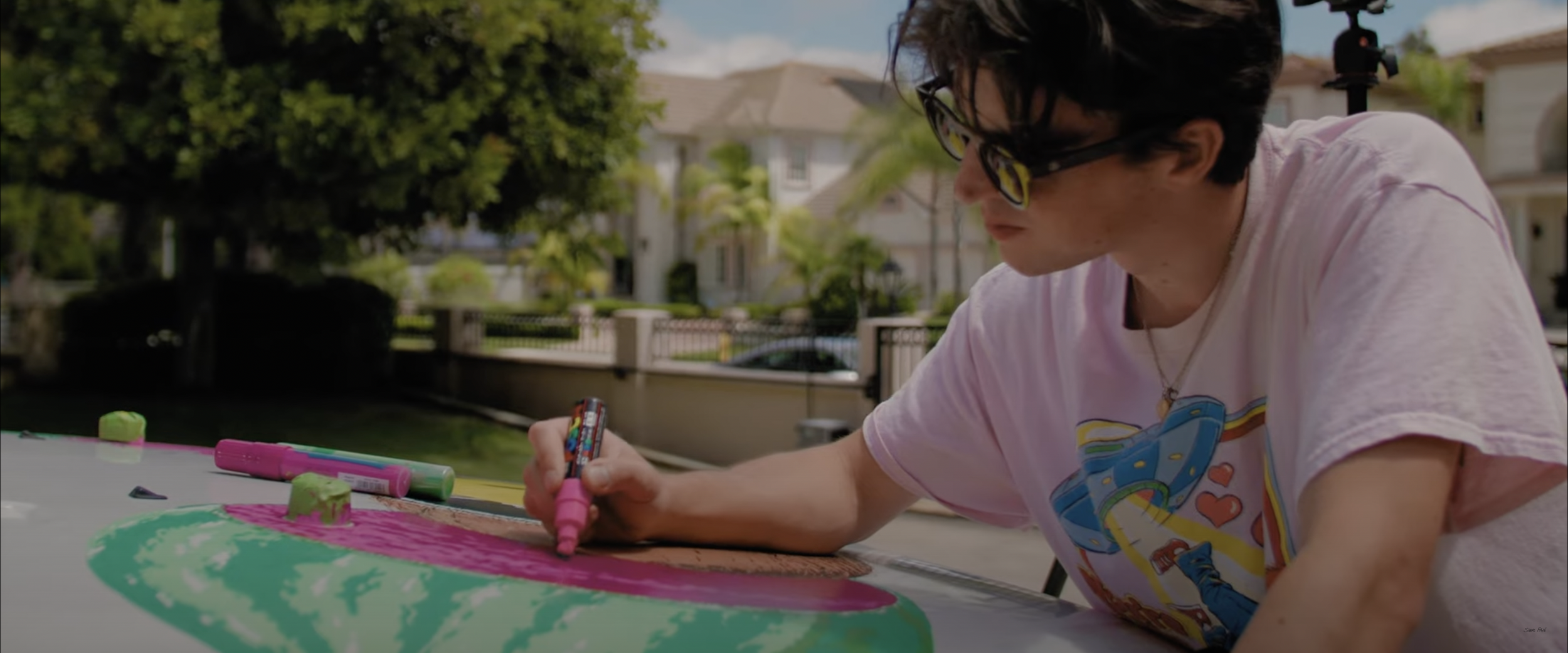 Person wearing sunglasses painting a design on a car with a pink marker, outdoors.
