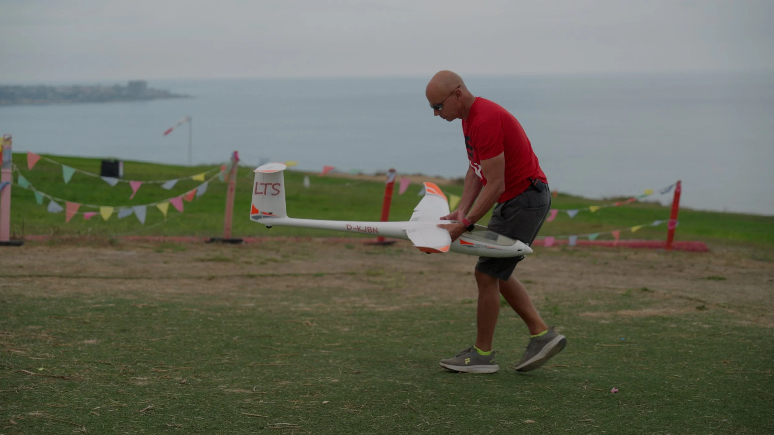 Person in red shirt holding a model glider plane near a coastal area with colorful flags in the background.