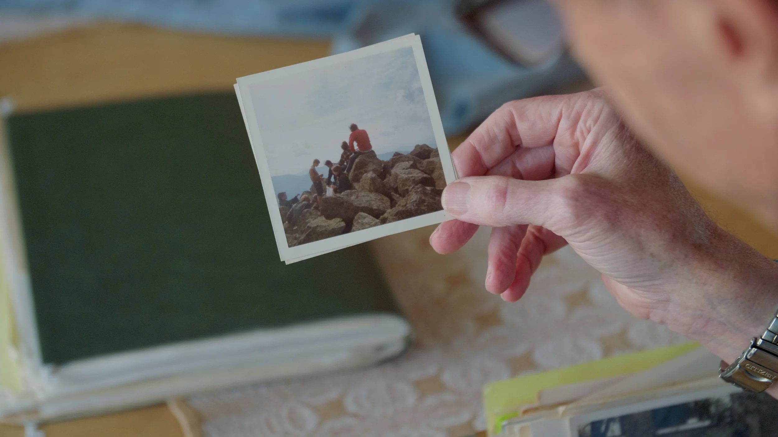 Person holding a vintage photo of people sitting on rocks, with a green photo album in the background.