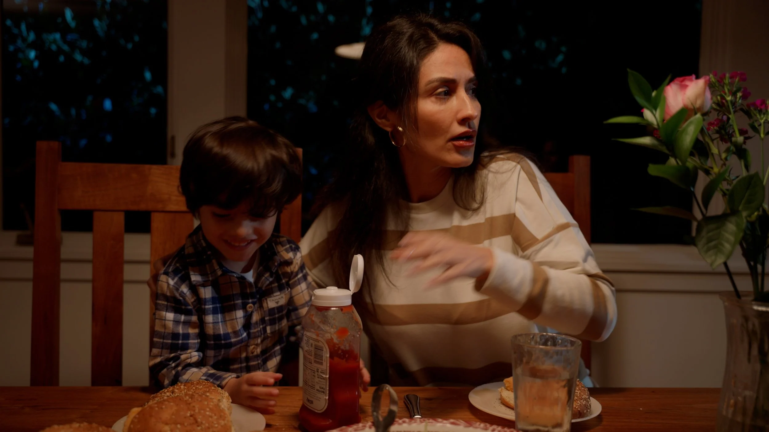 Woman and child sitting at a dinner table with burgers, ketchup bottle, and a glass of water; a flower vase is visible on the side.