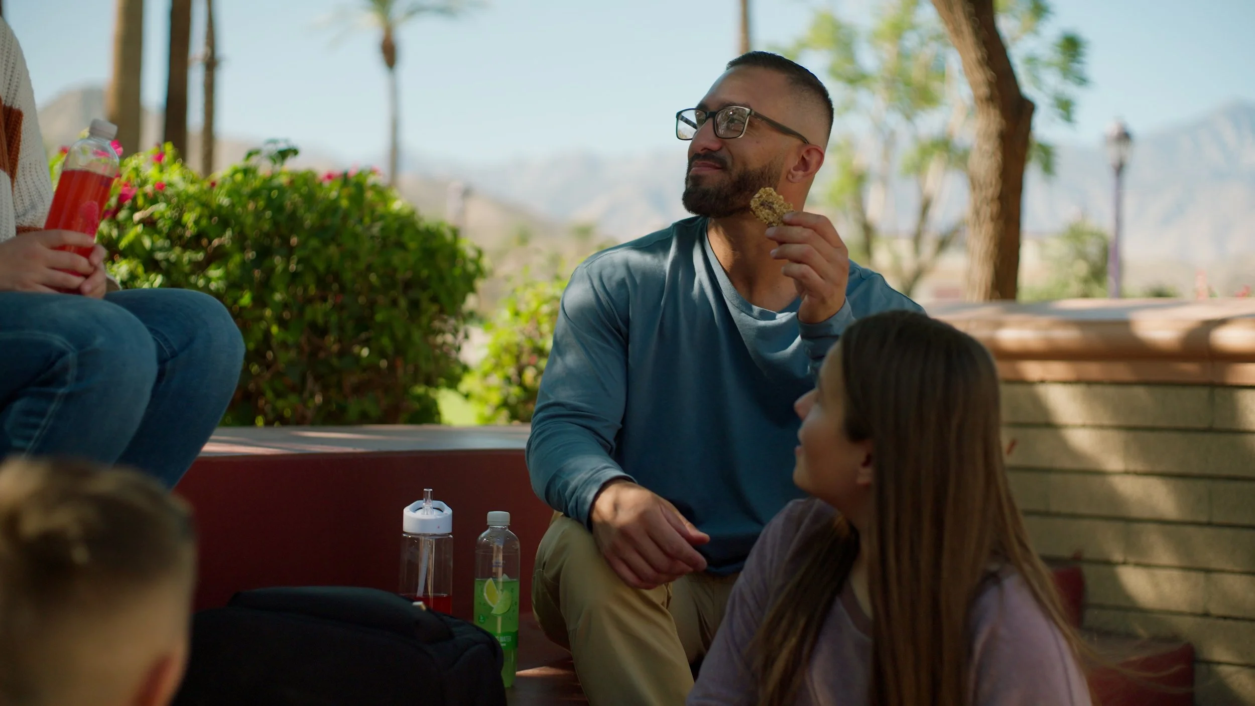 Group of people sitting outdoors, one holding a snack, two water bottles visible, green plants in background.