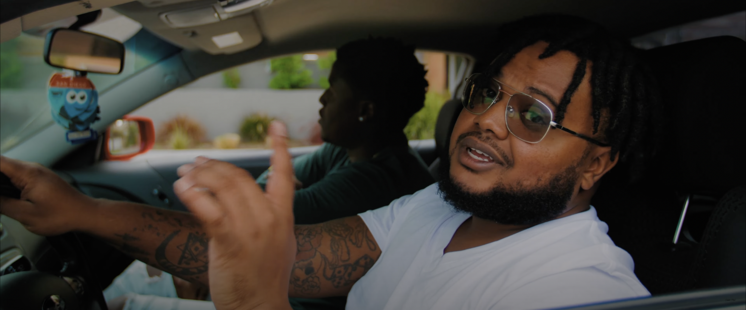 Two men seated in a car, one wearing glasses and pointing, with tattoos visible on his arm.