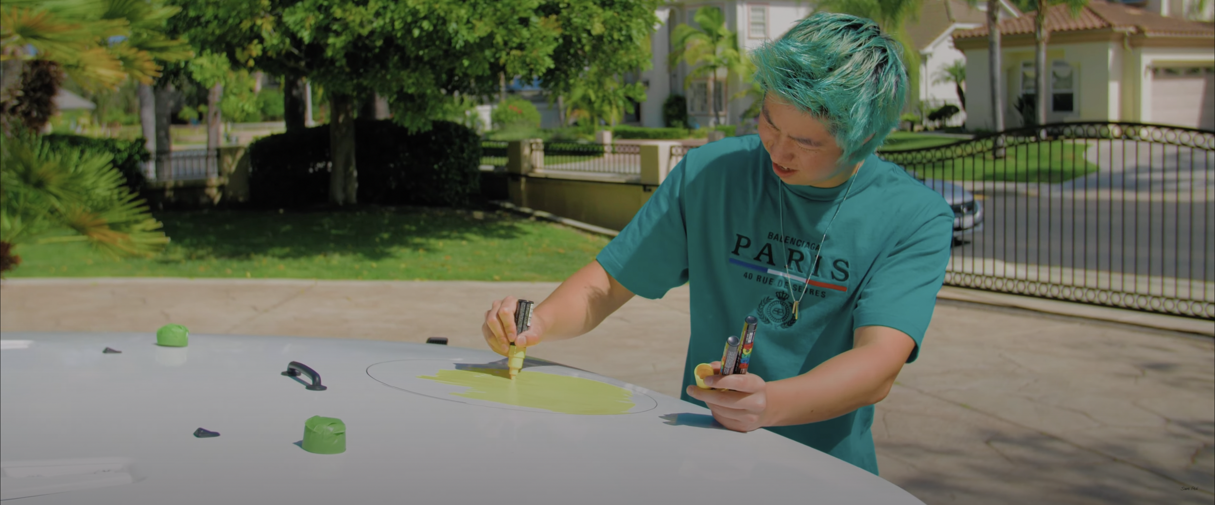 Person with turquoise hair painting on car hood outdoors