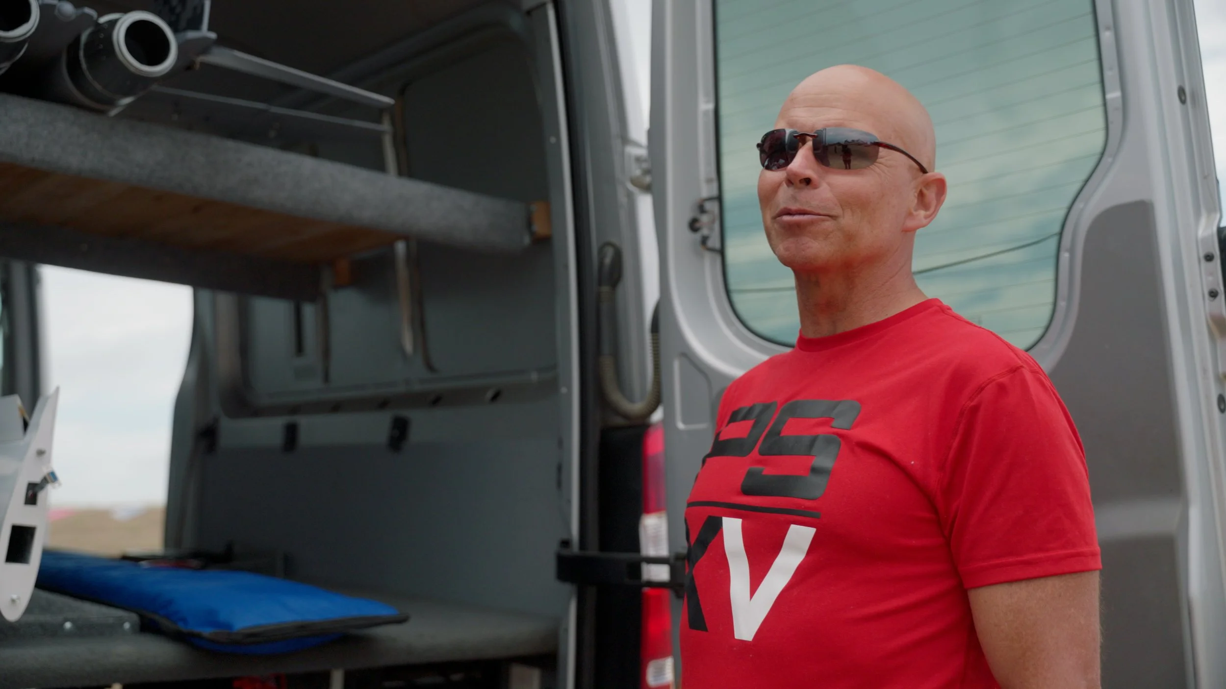 Man in sunglasses and red t-shirt standing beside an open van with interior shelves, holding model jet components.