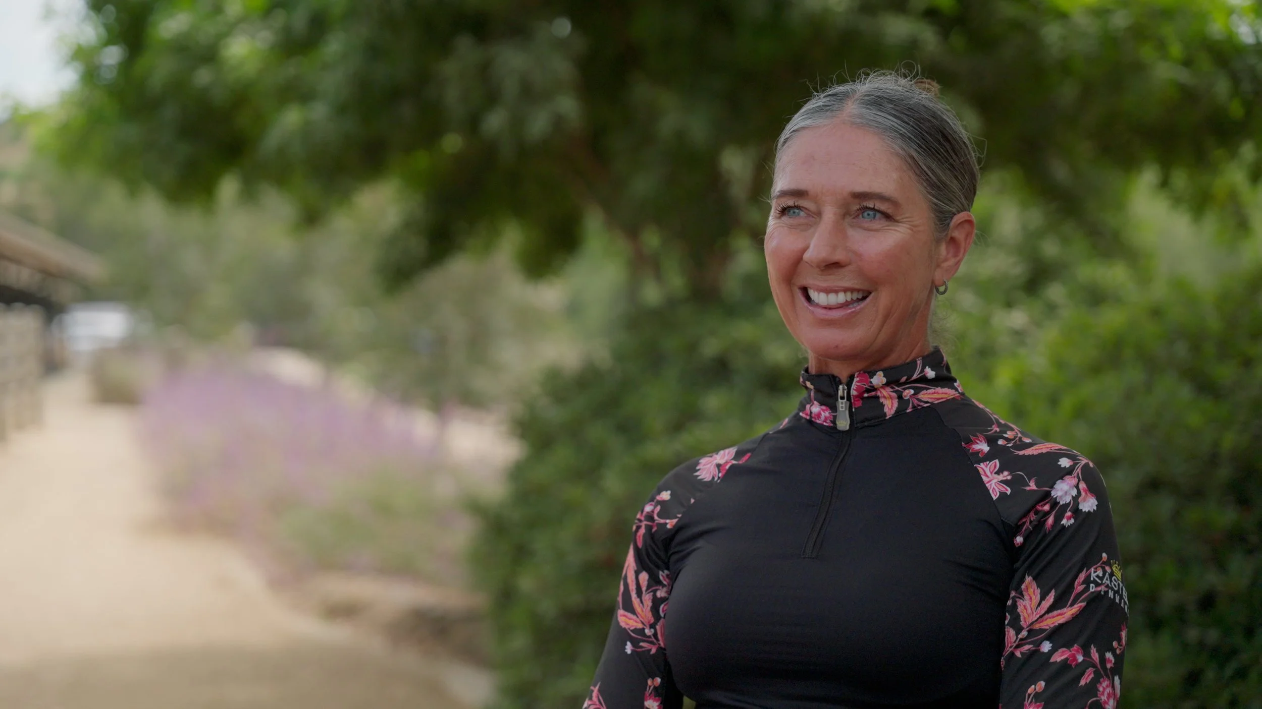 A woman smiling outdoors wearing a black floral top, with greenery in the background.
