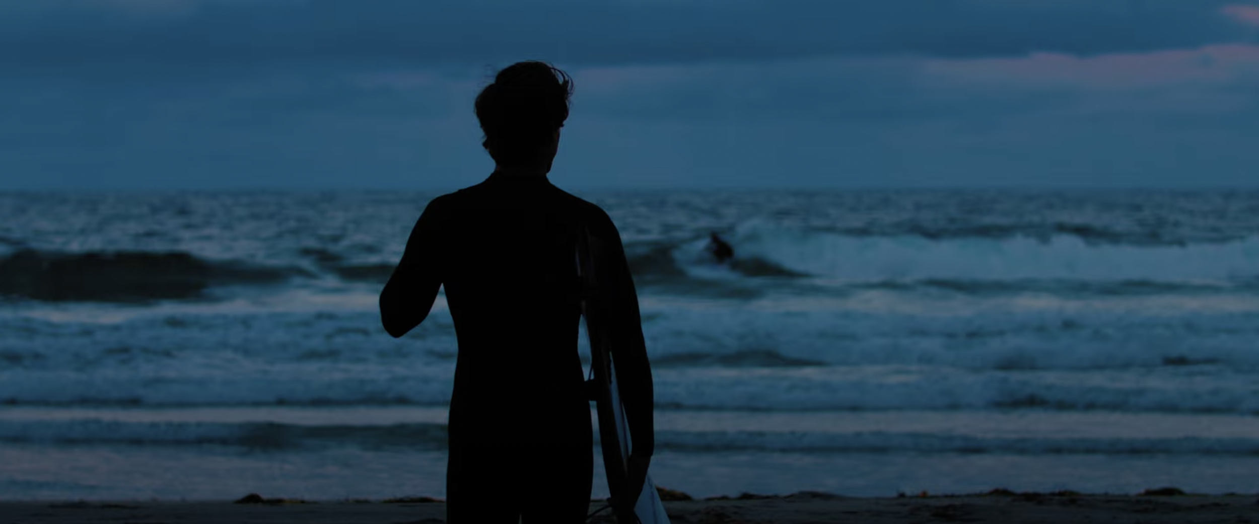Silhouette of a person with a surfboard facing the ocean at sunset with waves.