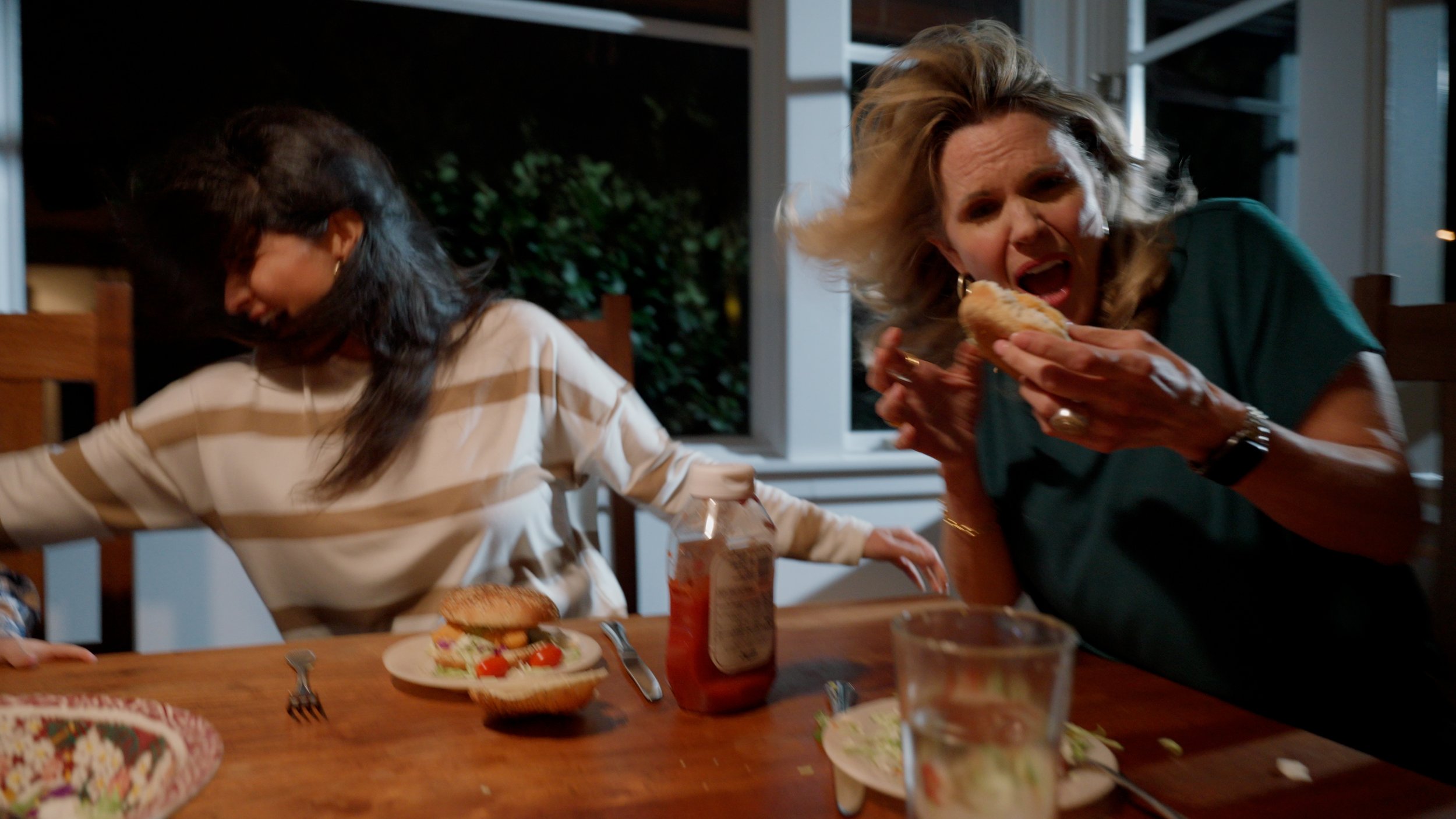 Two women at a dining table reacting to something off-screen, visibly startled, with food, including burgers and salad, on the table.