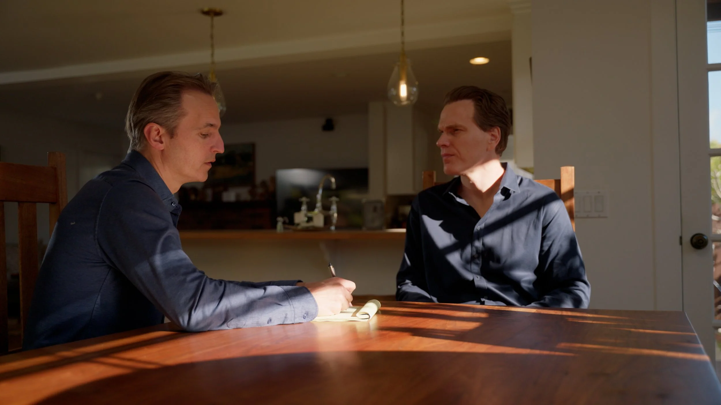Two men sitting at a wooden table, one writing on a notepad, indoors with soft lighting.