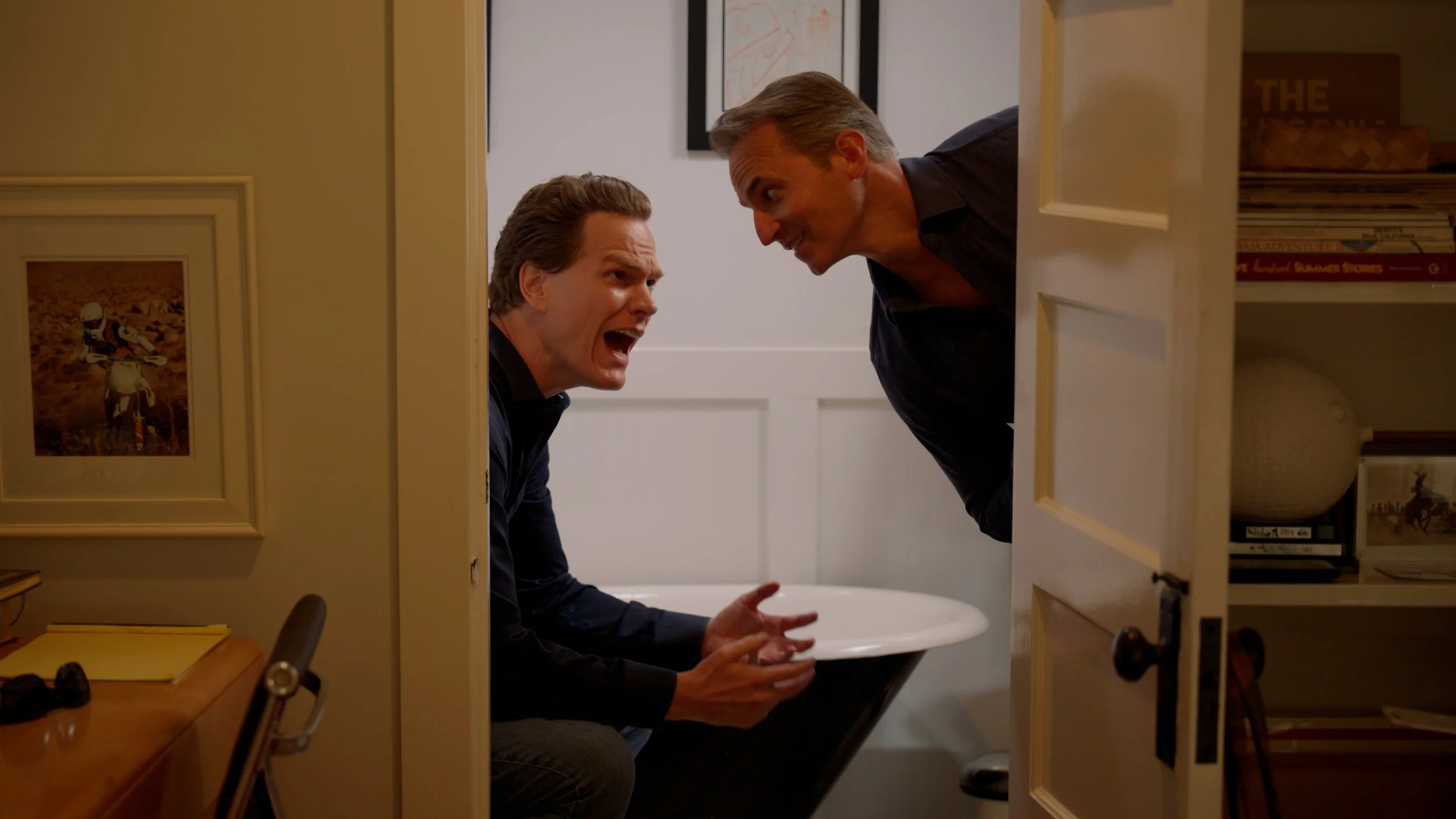Two men in a bathroom, with one sitting and the other leaning over a door, discussing something animatedly. A bathtub and framed pictures are visible.