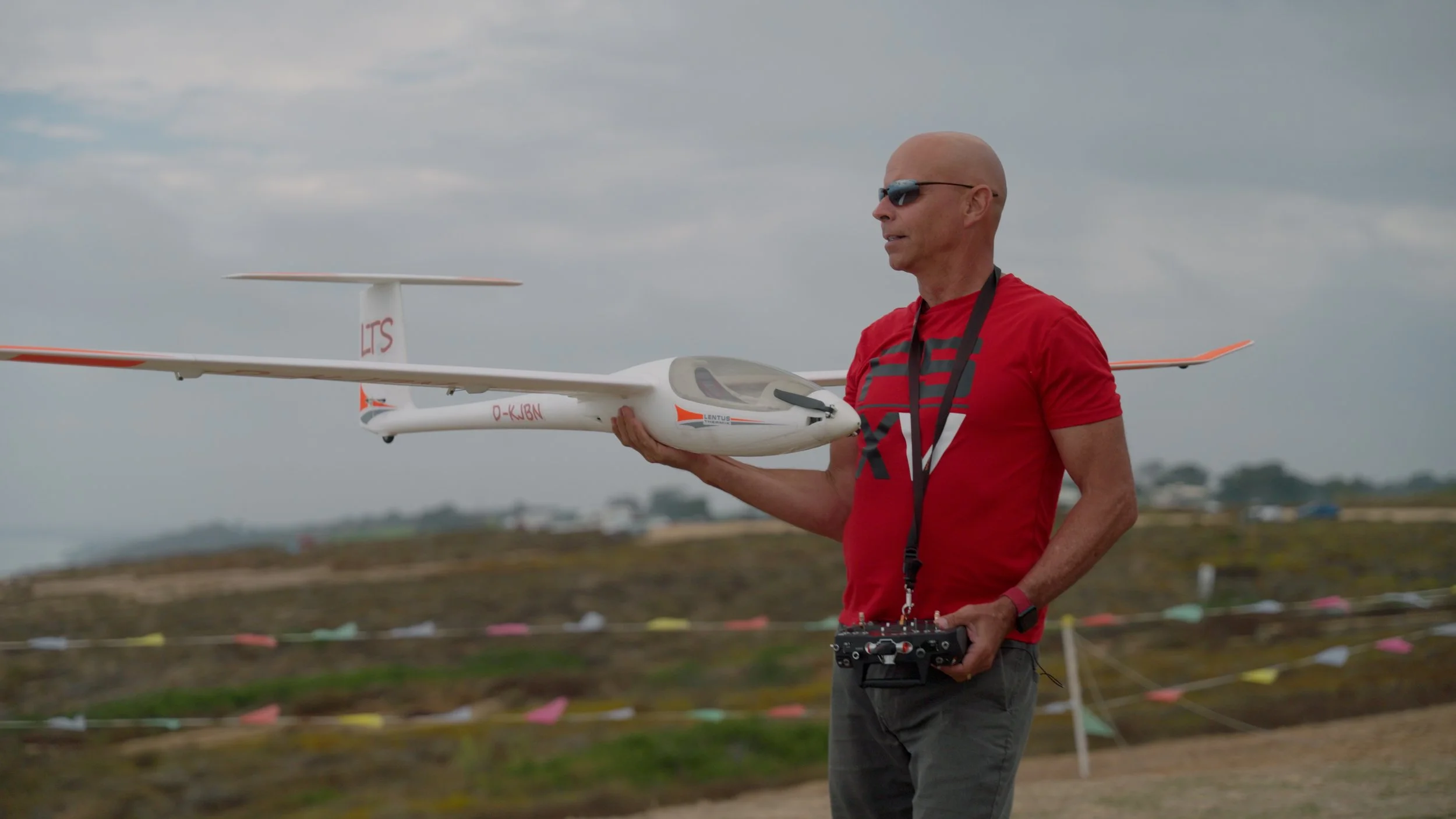 Person holding a large model glider airplane outdoors, wearing a red shirt, sunglasses, and holding a remote control.
