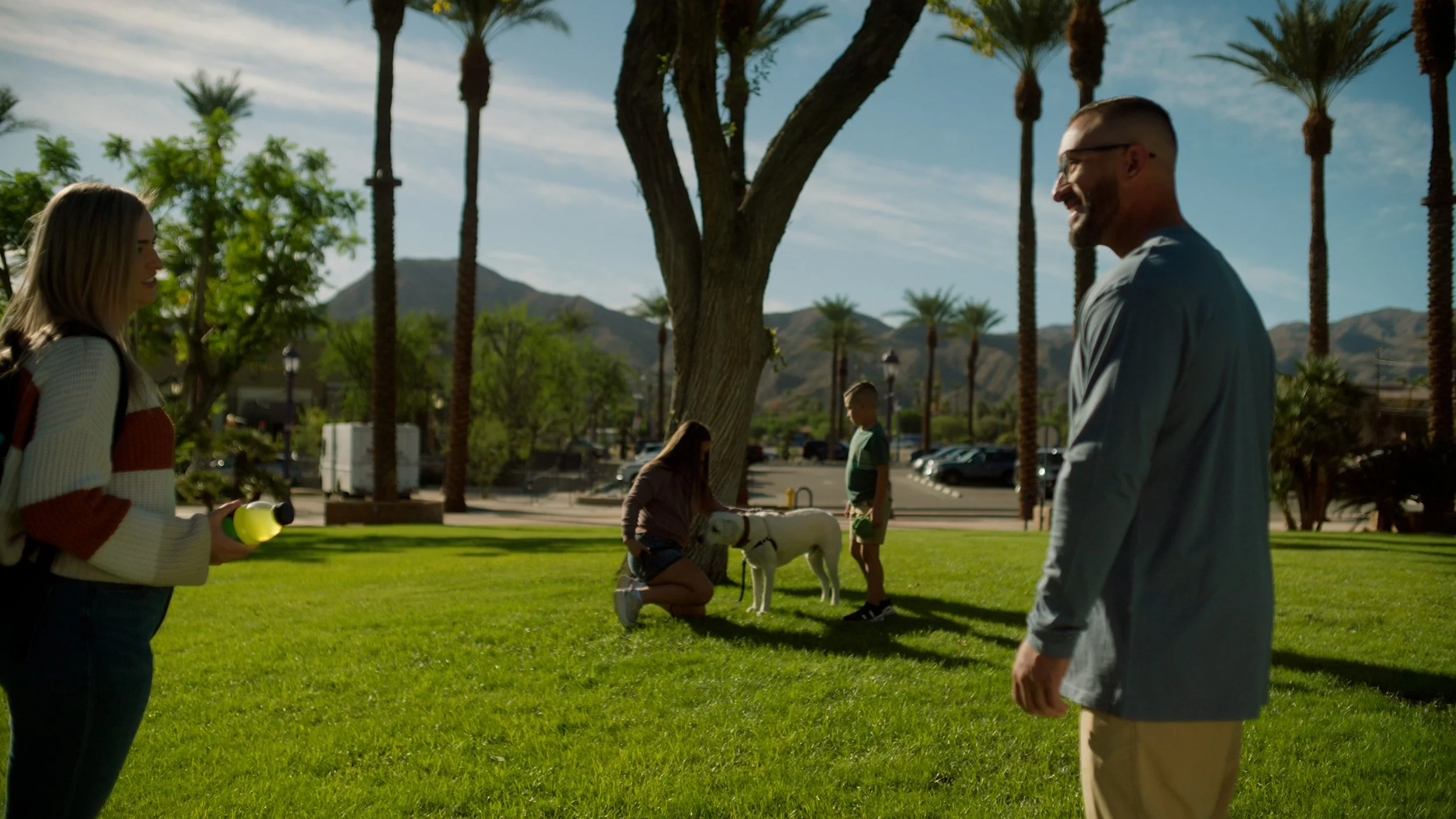 People conversing in a park with a dog, surrounded by palm trees and mountains in the background.